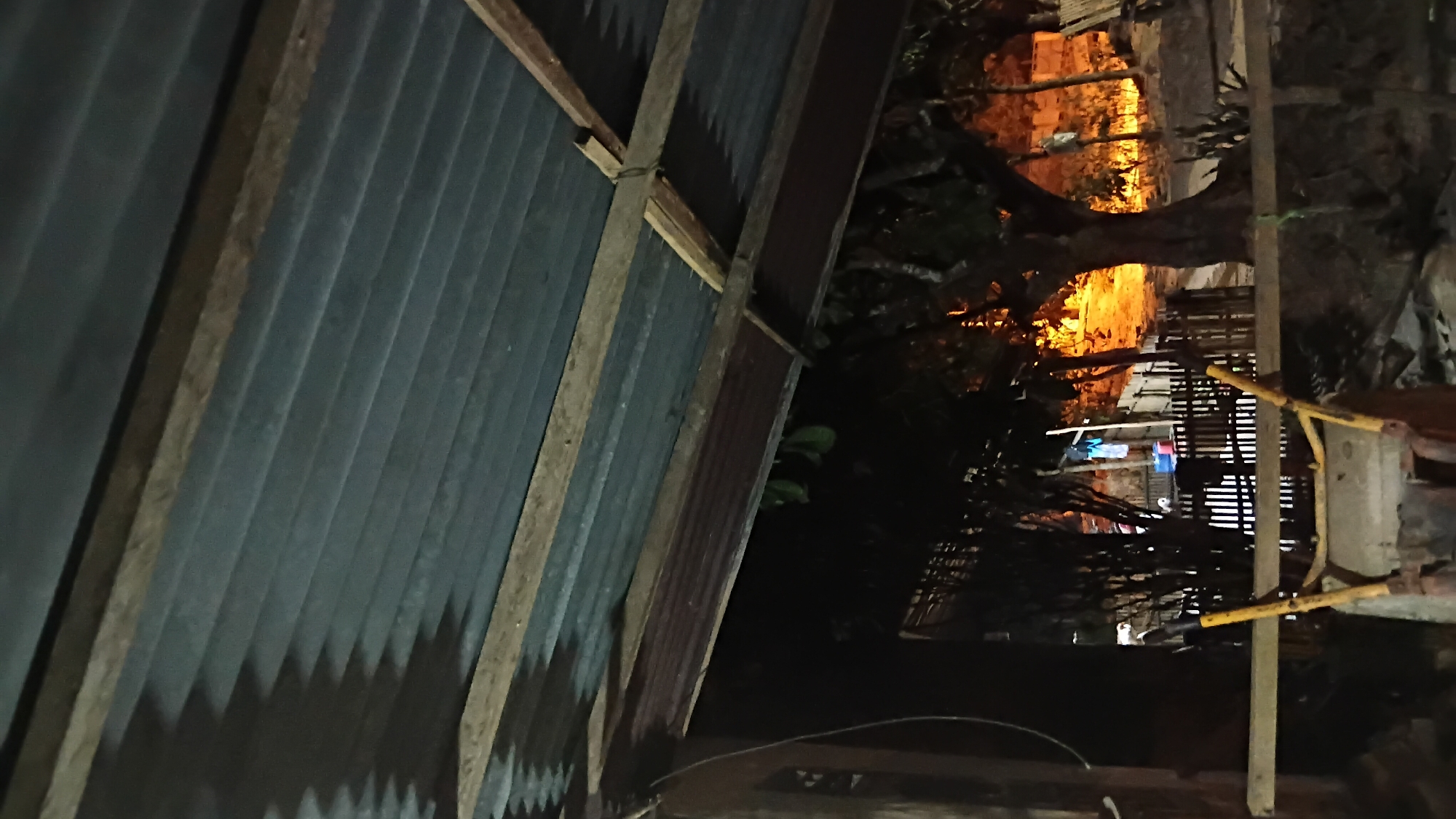 The image depicts an outdoor scene at night, likely in Prabumulih, Indonesia, under a corrugated metal roof supported by wooden beams. In the foreground, a yellow and brown wheelbarrow or similar garden tool is partially visible. Beyond this, a wooden fence encloses an area where laundry is hung on a line, with blue buckets visible. Trees are scattered throughout the scene, and a warm, orange glow from artificial lights illuminates the background, suggesting a residential or community area. The overall impression is one of a humble, functional outdoor space at dusk or night.