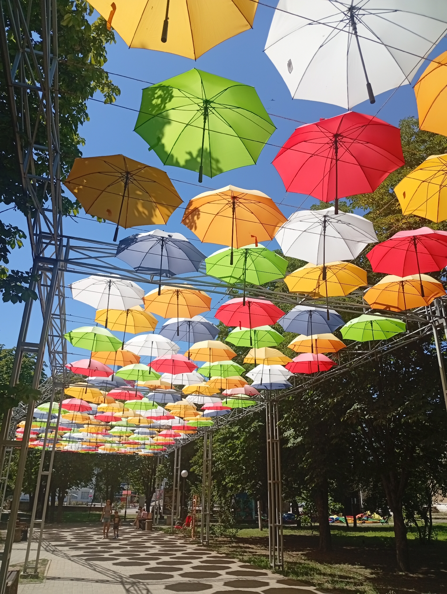 An overhead view captures a vibrant display of colorful umbrellas suspended by wires, forming a canopy over a pedestrian walkway in Pokrov, Ukraine. The umbrellas, in shades of yellow, green, red, white, and blue, are tightly packed, creating a visually striking effect against the clear blue sky. Sunlight filters through the umbrellas, casting circular patterns on the paved walkway below.
In the background, a metal structure, possibly part of a park or a temporary installation, supports the umbrellas. Lush green trees flank the walkway, and further back, buildings and park benches are visible. Two people, a woman and a child, are walking on the path in the foreground, their shadows elongating on the ground. Another group of people is seated on a bench in the middle distance. The scene suggests a lively outdoor public space, likely a park or a town square, decorated for an event or as a permanent art installation.