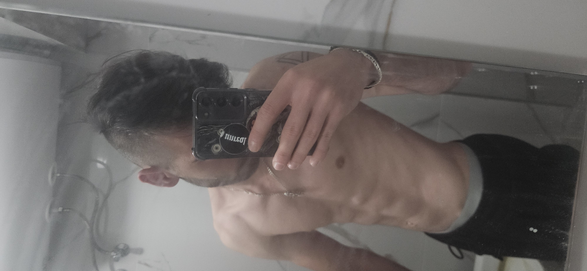 A selfie in a bathroom mirror shows a shirtless person with dark hair styled in a bun, wearing dark pants. They are holding a smartphone with a black case adorned with a round sticker. The background features a marble-patterned wall and glimpses of bathroom fixtures. There is no evidence to suggest the location is in Plovdiv, Bulgaria.