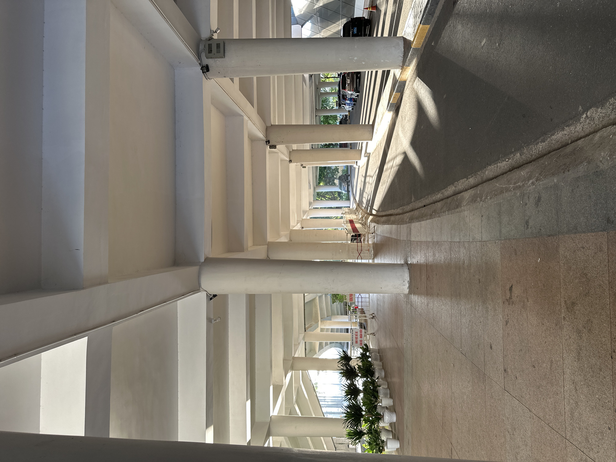 The image shows a view from under a large, white, multi-story concrete structure, likely a parking garage or covered walkway, in Da Nang, Vietnam. White circular columns support the structure, casting long shadows across an asphalt road on the right and a tiled pedestrian walkway on the left. A line of potted green plants with dark leaves is arranged along the walkway. In the background, several cars are parked under the structure, and trees are visible in the distance. The lighting suggests it is either early morning or late afternoon, with sunlight streaming in from the right, creating a strong contrast between light and shadow. A small orange traffic cone is visible near the road.