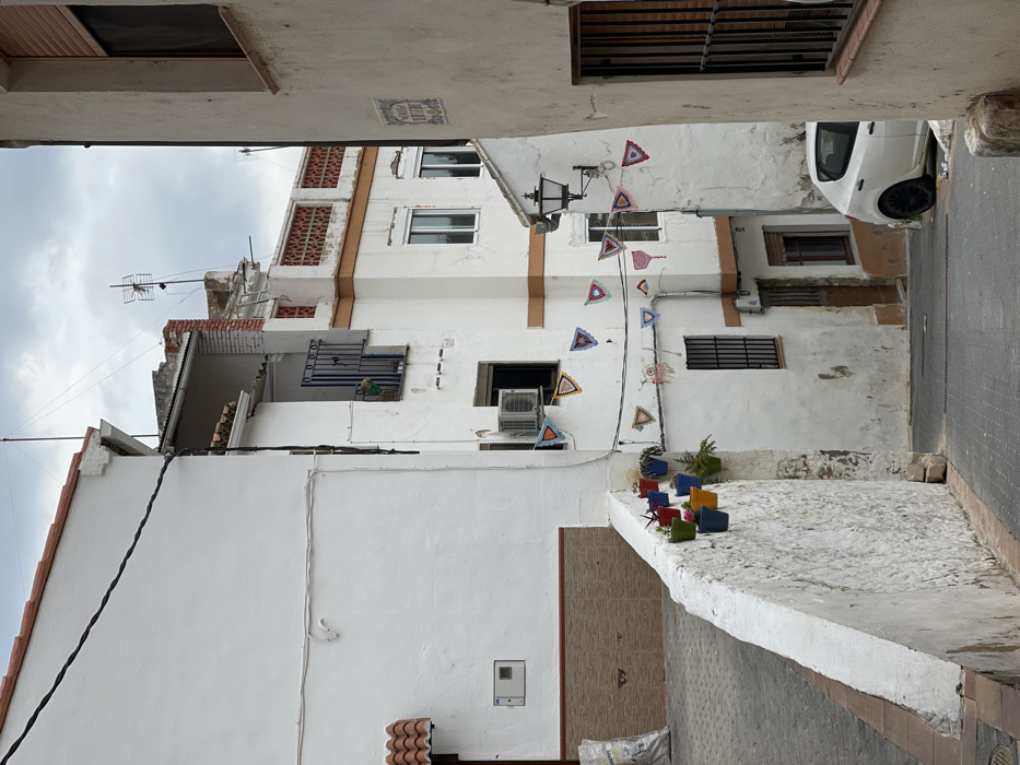 This image depicts a narrow, sloping street in Buñol, Spain, characterized by closely packed, predominantly white stucco buildings under an overcast sky.
In the foreground on the left, a whitewashed, sloped stone or concrete retaining wall runs parallel to the viewer, adorned along its top edge with a series of approximately nine brightly colored plastic flower pots (red, green, blue, yellow, purple, black, teal) containing small green plants. Behind this wall, an upward-sloping paved area is visible, leading to the base of a building where a large white sack (possibly containing construction material) rests. The leftmost building features a clean white facade with a red-tiled roofline and a lower section clad with light brown tiles. A dark electrical cable stretches across its upper wall horizontally, then vertically downwards, and a white utility box is mounted on its lower section.
The street itself is paved with dark grey asphalt. Towards the center, multiple white buildings line the descending street. One prominent building on the left features a second-story balcony with blue vertical bars and a potted plant. Below this, an open window has an external air conditioning unit mounted above it. A string of colorful, triangular, crochet-style pennants or bunting, some with circular, dreamcatcher-like patterns, is strung across the street, attaching to this building and extending to the right. Further down the street, another window with dark vertical bars is visible on the same building, alongside a dark brown wooden door.
On the right side of the street, a light-colored building with a textured facade features a window with dark vertical bars. A ceramic tiled street sign or plaque with blue lettering is affixed to its wall. An ornate, dark-colored street lamp with a frosted glass globe is mounted on a building wall further back on the right. Above this, a building exhibits a decorative terracotta-colored lattice pattern along its rooftop, and various rooftop antennae are visible against the grey sky.
A white compact car, possibly a SEAT Ibiza, with black wheels is parked on the right side of the descending street, facing away from the viewer, partially obscured by the foreground building on the right. No people are visible within the scene. The overall impression is a quiet, residential urban environment.