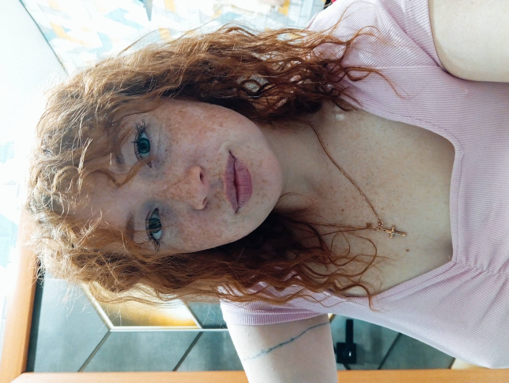 A close-up, portrait-style photograph shows a person with red, curly hair and freckles wearing a light pink, short-sleeved shirt. The person is also wearing a gold necklace with a cross pendant. A tattoo is visible on the person's left arm. The background appears to be an interior setting with geometric patterns and a light source creating highlights. Location: Yurgamysh, Russia cannot be confirmed.