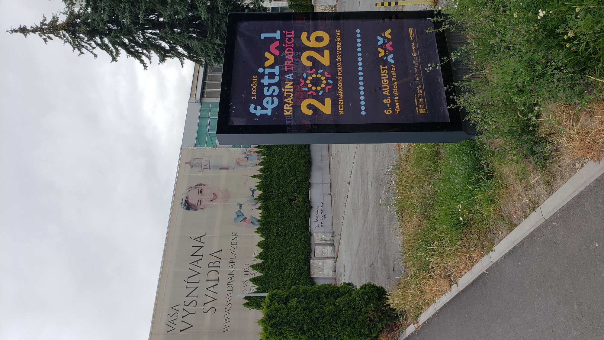 In the city of Prešov, Slovakia, an outdoor digital billboard displays information about the 1st annual "Festival Krajín a Tradícií" (Festival of Countries and Traditions). The festival is a celebration of international folklore and will take place from August 6th to 8th on Hlavná ulica in Prešov. The billboard is positioned in front of a large building with a banner that reads "Vaša VYSNÍVANÁ SVADBA" (Your Dream Wedding) and a website address. In the background, a tall evergreen tree stands to the right of the building, and a row of manicured green bushes lines the area in front of the building. The foreground shows a paved sidewalk and a grassy area with some weeds and wildflowers.