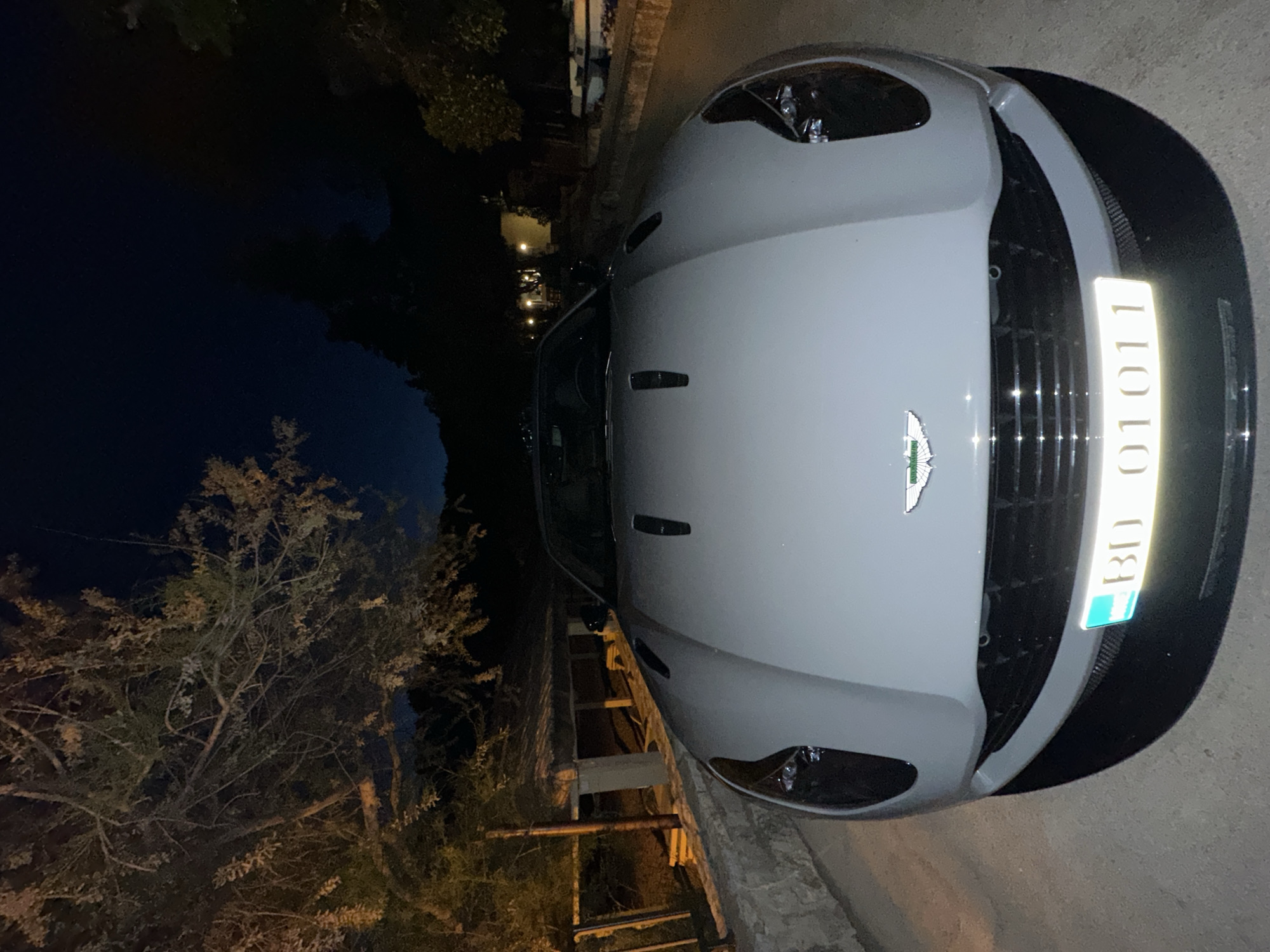 The image shows the front of a white Aston Martin parked on a street. The license plate reads "BD 01011." The vehicle is parked in a residential area, with trees and buildings visible in the background. The photo was taken at night in Petrovac centar, Montenegro.