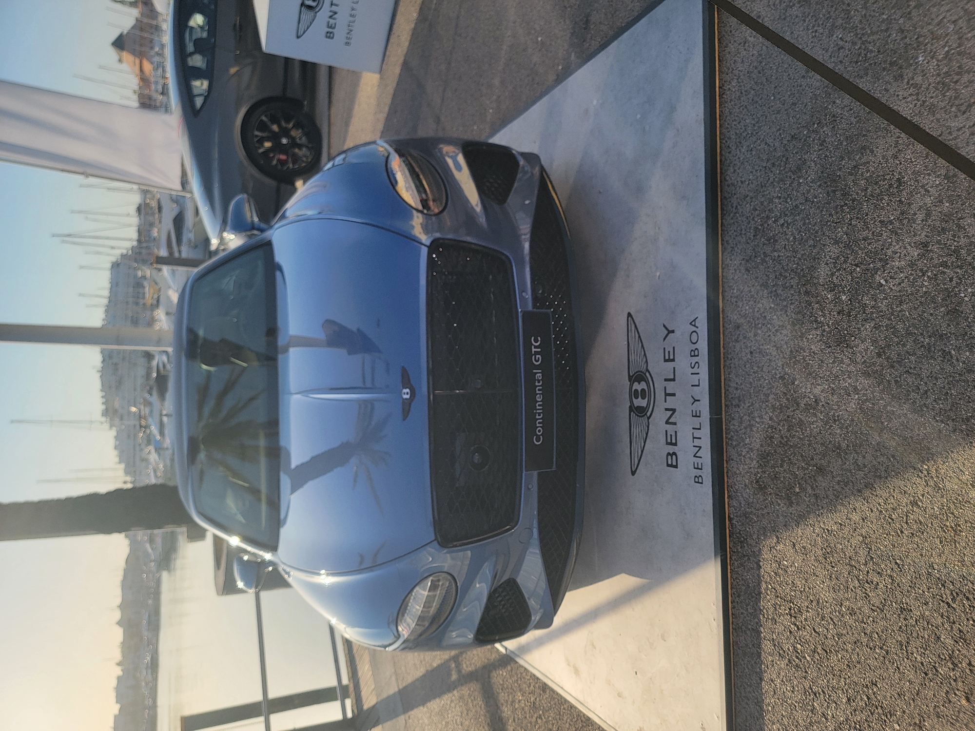 A blue Bentley Continental GTC is parked on a raised platform at a waterside location in Quarteira, Portugal. The car is positioned in the foreground, facing the camera. Behind the car, a gray Bentley coupe is parked to the right, and a white banner is visible. The background shows a marina filled with sailboats and buildings. The overall lighting suggests it is either sunrise or sunset, with the warm glow of the sun. The platform the blue Bentley is on has the Bentley logo and "BENTLEY LISBOA" printed on it.
