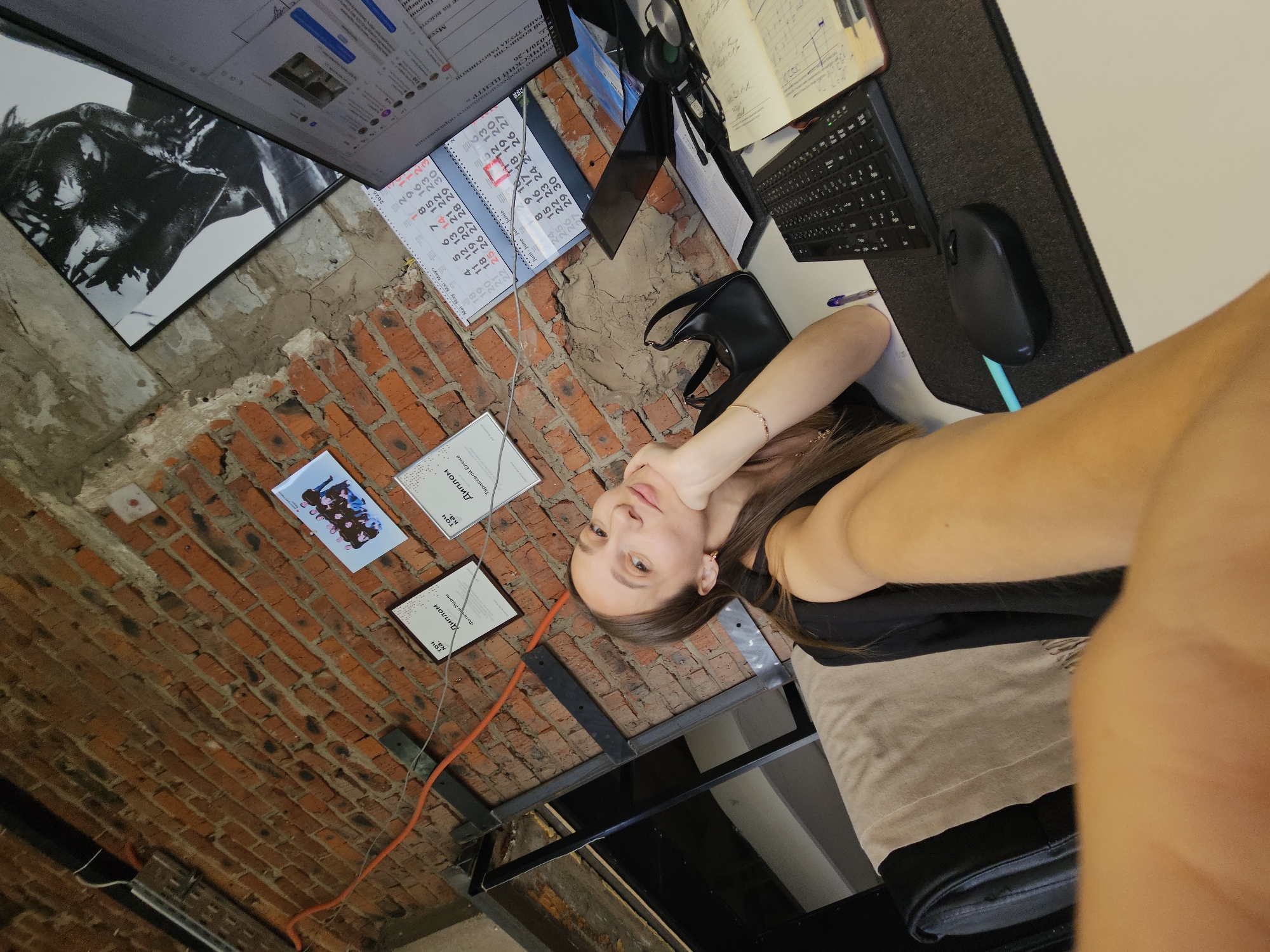 A young woman is captured in a selfie, taken from a slightly high angle, in what appears to be an office or creative workspace in Chaikovsky, Russia. She has long brown hair and is looking directly at the camera with a calm expression, resting her chin on her right hand. She is wearing a dark top.
The background reveals an exposed red brick wall with sections of concrete. Her workstation includes a desk with a computer monitor displaying a Russian-language web page or application, a keyboard, a black mouse on a mousepad, and headphones. An open notebook with handwritten notes and a black handbag are also on the desk. A desk calendar shows multiple months, including "Mai May Mal Mayo" and "Juni June Jun Junio," with some dates highlighted, a visible year "2026," and a prominent number "9162330."
On the wall, several items are hung: a large, framed black-and-white artwork depicting a person's head and shoulders, a smaller picture of a group of people (likely 4-5 individuals), and several framed "Диплом" (Diploma) certificates. Visible names on the diplomas include "Фотин Марии" and "Тарасовой Елене." An orange electrical cable runs along the brickwork, and metal structural elements are also present, contributing to an industrial aesthetic. Text visible on the screen and documents includes "ДИЧЕСКИЙ ЦЕНТР," "РАНЫ ТРУДА РАБОТ" (possibly "Labor Protection"), "Муса," "на высоте," and fragments like "Причи," "вері," "комиссии," and "О-2020/1-26."