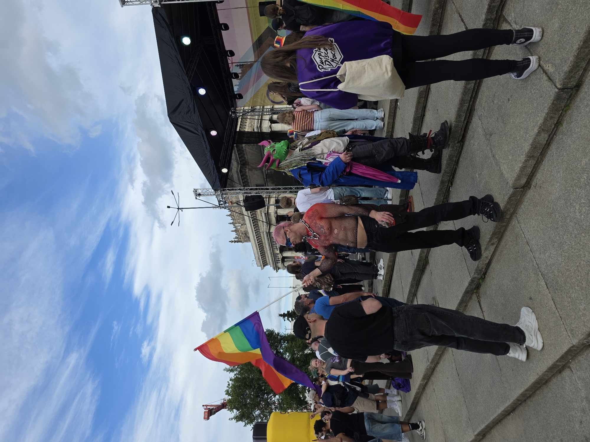 The image shows a crowd of people on a wide, outdoor staircase in front of a building with classical architecture. A stage with a black awning is set up in the background. Several people are holding or displaying rainbow flags, indicating a Pride event.
One individual in the foreground is wearing a sheer, red, mesh top, black pants, and a chain necklace. They are holding a rainbow flag on a pole. To their right, another person is wearing a green dragon mask, a blue coat, and black pants. Further to the right, a person in a purple jacket with "Purple Disco Machine" embroidered on the back is standing with a canvas tote bag. Other individuals in the crowd are dressed in casual attire.
In the background, a large, yellow cylindrical object is visible, as is a red construction crane. The sky is partly cloudy with blue skies. The building in the background appears to be the Palace of Culture and Science, a prominent landmark in Warsaw, Poland. The event seems to be taking place in front of this building, likely on the plaza or surrounding area. The overall atmosphere suggests a public gathering or celebration.