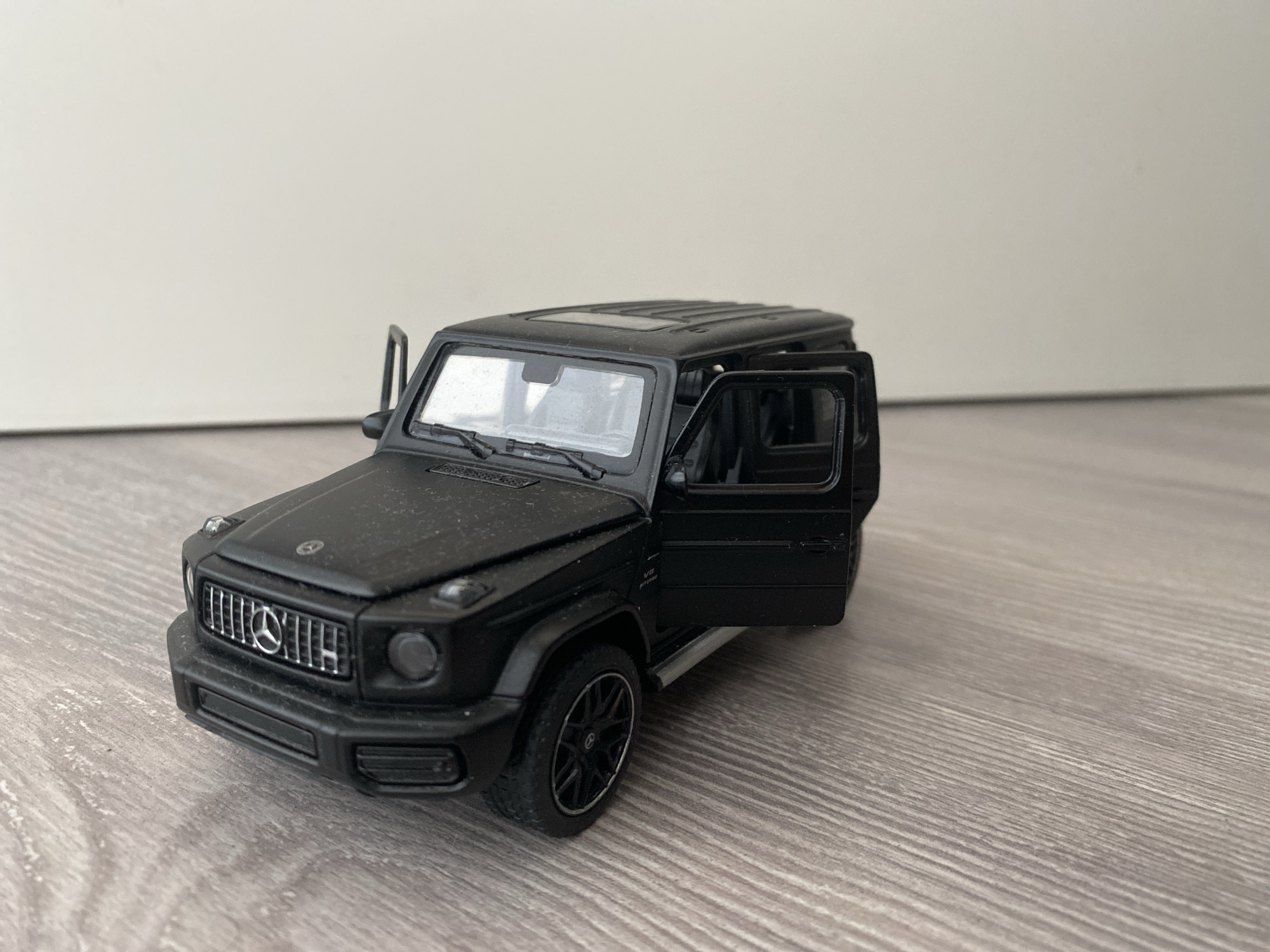 A black Mercedes-Benz G-Class toy car is positioned on a wooden floor, with its driver's side door open. The toy car is detailed, featuring a grille with the Mercedes-Benz logo, visible headlights, and the "V8 BITURBO" badging on the side. The car appears to be covered in a light layer of dust, suggesting it has been in storage or on display for some time. The background consists of a plain, off-white wall. This scene is not located in the city of Łódź, Poland; it is a generic indoor setting.