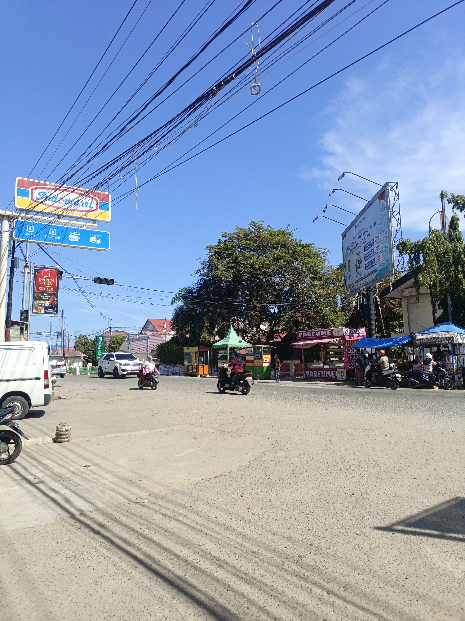 This is a medium shot, eye-level, outdoor, color photograph taken on a bright, sunny day. The scene depicts a street intersection in a semi-urban area, likely Mesjid Tuha, Indonesia. Overhead, a dense network of power lines crisscrosses the clear blue sky, casting long, distinct shadows on the paved road below.
The foreground is dominated by the road and its surface. To the left, a white van is parked, with the front of a motorcycle visible. Further into the intersection, a white SUV is seen, with a person on a motorcycle in front of it. In the mid-ground, several small roadside stalls are set up. One stall, pink in color, is adorned with "PARFUME" signage, with prices displayed. Another stall, under a blue awning, also appears to be selling goods, with several people gathered around.
Above the intersection, several signs are visible. A prominent "Indomaret" sign, a popular convenience store chain, is mounted on a pole, accompanied by smaller signs indicating "pulsa", "payment", "DEBIT", and "PAKET". A traffic light is also visible, although its current state is unclear. A large billboard with text and imagery, possibly an advertisement, is mounted on a metal frame to the right of the intersection.
The overall atmosphere is one of everyday activity. The bright sunlight and clear sky suggest it's daytime. The presence of multiple motorcycles, stalls, and people indicates a bustling commercial area. The visible text confirms the location is in Indonesia, with references to local brands and services.