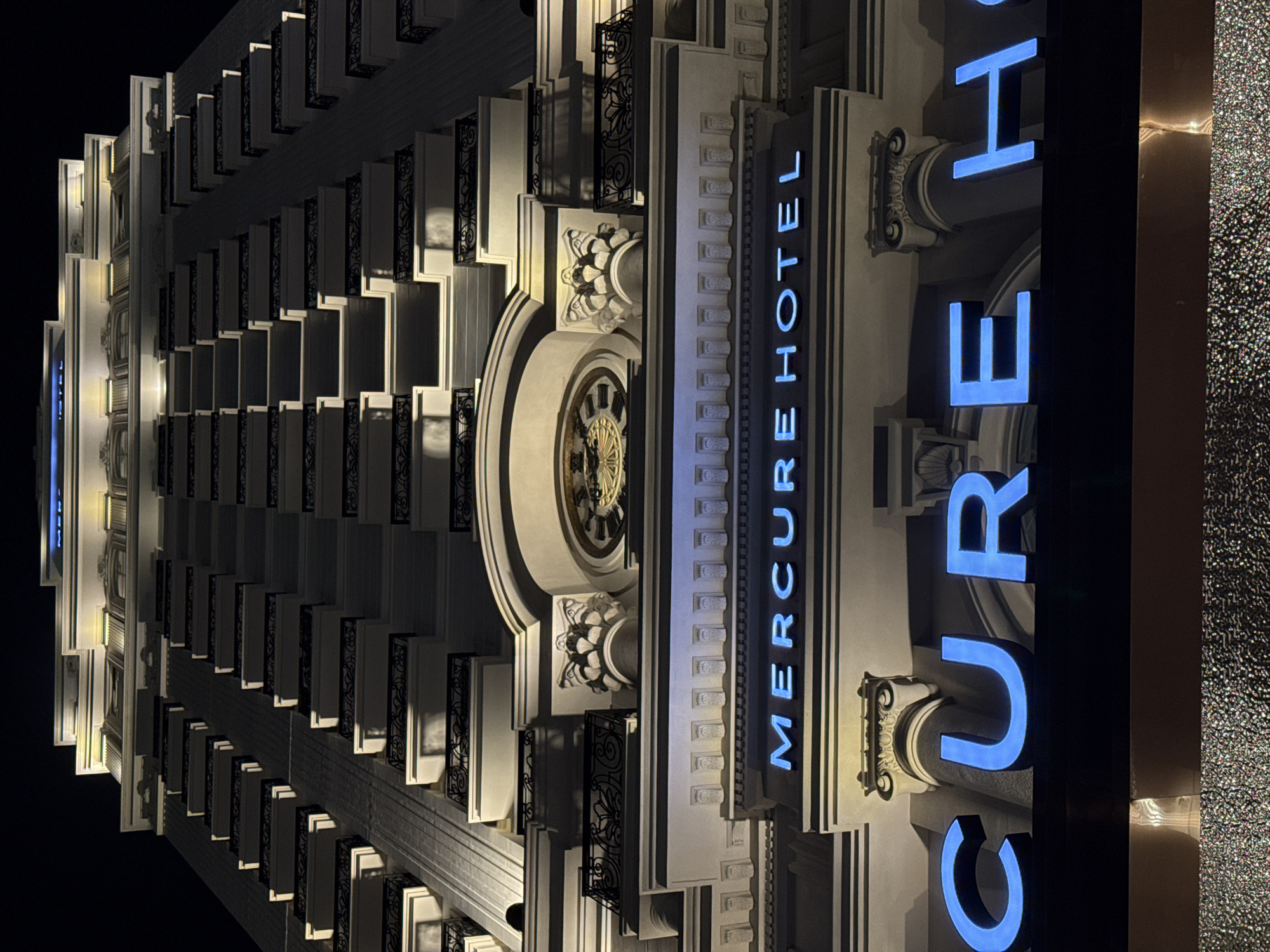 The image displays the illuminated facade of the Mercure Hotel at night in Nha Trang, Vietnam. The multi-story building features elegant, classical-inspired architecture with repeating patterns of recessed windows or balconies, each adorned with ornate black metal railings. Warm yellow lights highlight the white or light-colored stone facade, emphasizing its intricate cornices, moldings, and decorative carvings, including stylized floral motifs and capitals.
A prominent blue neon sign, oriented vertically in the image, spells out "MERCURE HOTEL" along a section of the facade. This sign is integrated into the building's design, flanked by architectural details. A large, ornate circular element resembling a clock face with intricate golden accents is visible, positioned near the "HOTEL" portion of the sign. The dark night sky provides a stark contrast to the brightly lit building. The overall scene conveys a sense of grandeur and sophistication of the hotel's exterior during the evening hours.