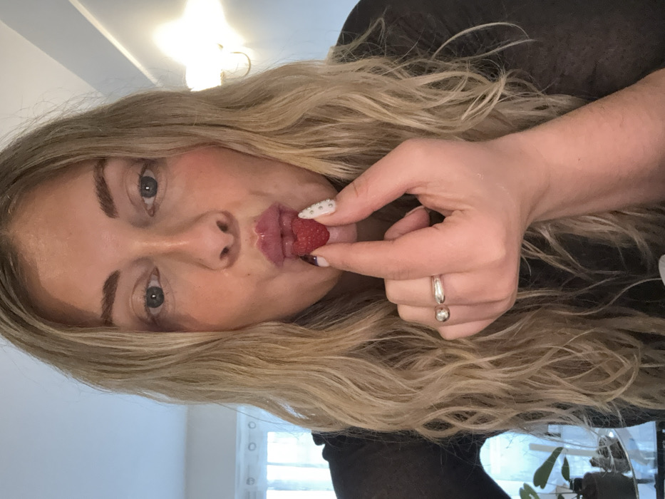 This image depicts an adult female subject in an indoor residential setting, identified as being in the city of Paris, France. The subject, with long, wavy blonde hair and blue-grey eyes, is looking directly at the camera. She holds a single red raspberry between her left thumb and index finger, positioned directly in front of her pursed, glossy lips. A silver-colored ring is present on her left ring finger. Her left index fingernail features white polish with small embedded studs, contrasting with a darker polish on her thumb. She is wearing a dark, possibly sheer garment. The background is out of focus, showing a window with sheer white curtains on the left and an illuminated wall sconce on the right. A potted plant is partially visible on a surface in the lower-left background.