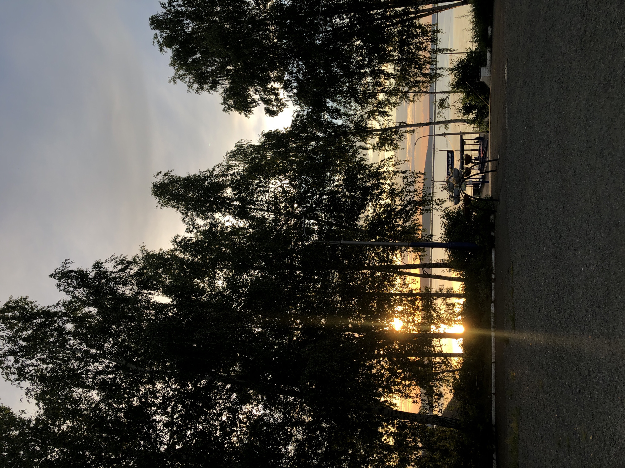 The image shows a silhouetted view of trees against a sunset over the Kama River in Chaikovsky, Russia. The foreground is a gravel surface. Behind the trees, the river is visible, with a faint glow from the setting sun. A pier or a small structure with some people on it can be seen on the riverbank. The sky is partly cloudy with a warm, soft light.