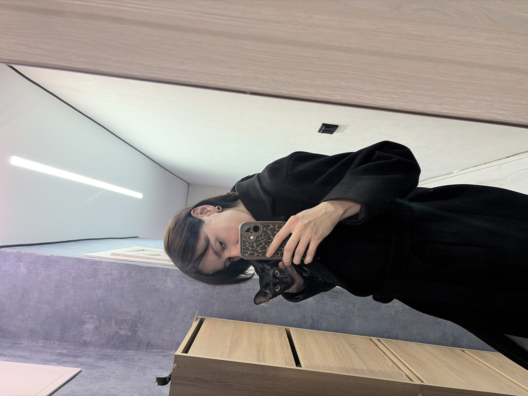 A woman wearing a black coat takes a selfie in a mirror with a black cat. The phone has a leopard-print case. Behind the woman is a wooden cabinet. The room has concrete walls and a white ceiling with recessed lighting. The location is Chaikovsky, Russia.