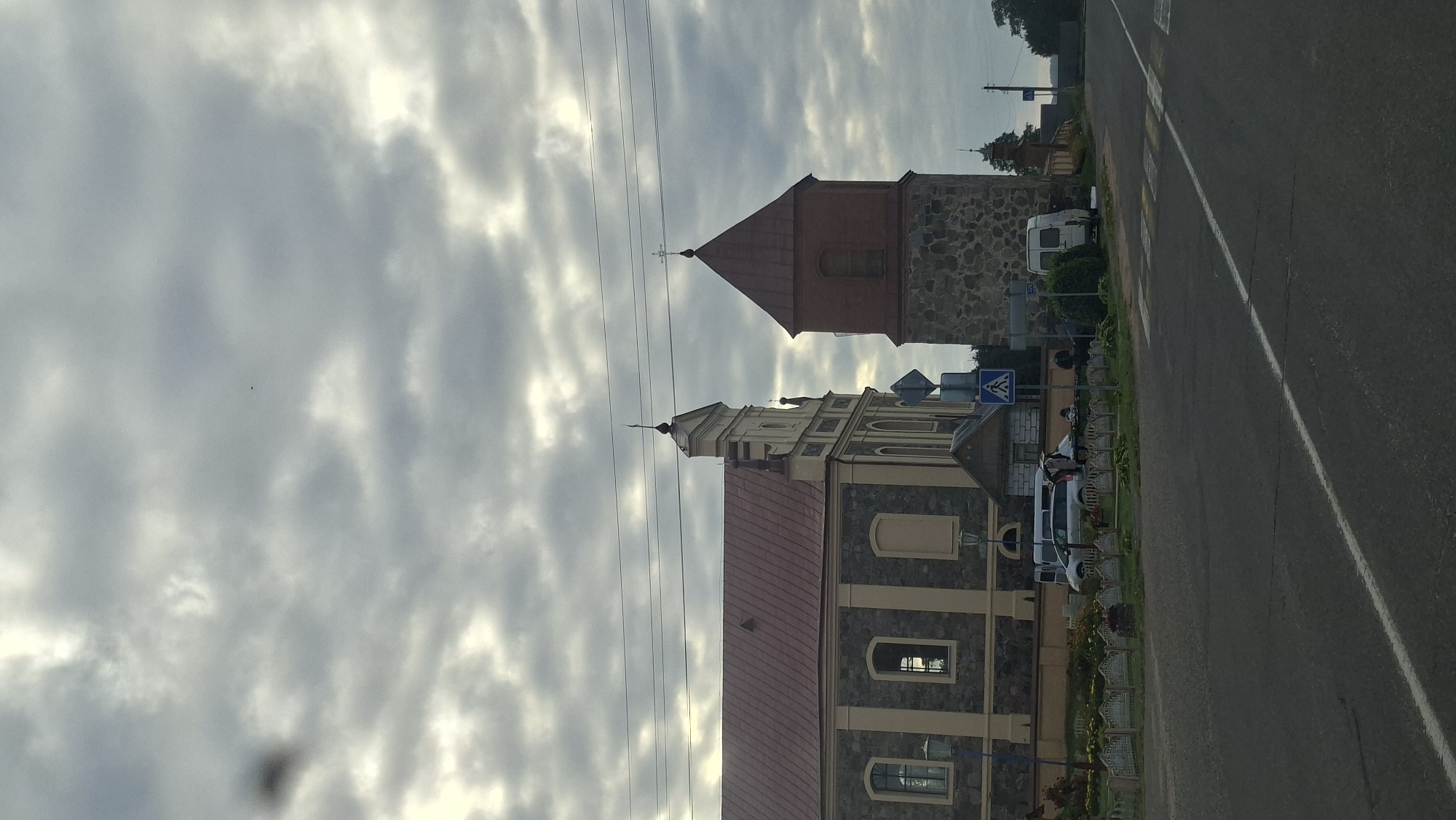 The image depicts a street scene in the city of Валкалацкі сельскі Савет, Belarus, under a cloudy sky. On the left, a large stone and stucco building with a red-tiled roof, likely a church, is visible. Adjacent to it on the right is a stone and brick bell tower with a red, pyramid-shaped roof topped with a cross. Several vehicles, including a white minivan and a silver car, are parked near the church. People are gathered near the vehicles, some appearing to be interacting. A pedestrian crossing is marked on the asphalt road in the foreground. The environment appears to be a town or village setting with some greenery and fences visible.