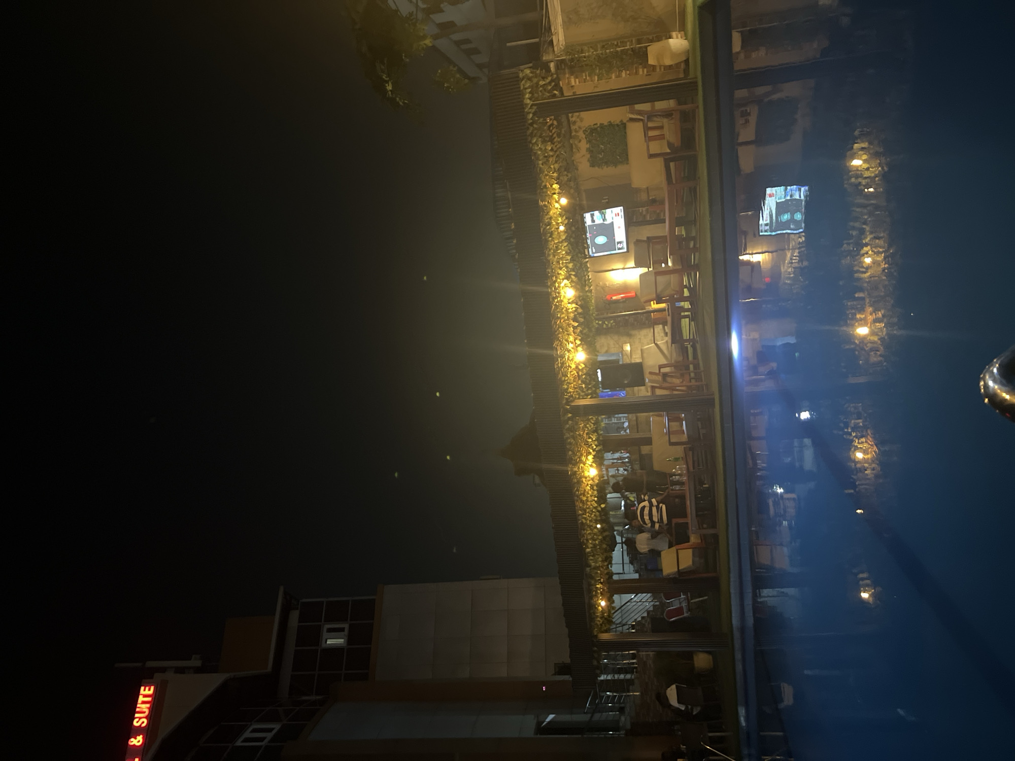 The image depicts a nighttime scene, likely an urban leisure area in Abuja, Nigeria, featuring a brightly lit establishment reflected on a calm, dark surface.
The main focus is a multi-level structure, possibly a restaurant or lounge, adorned with warm string lights and decorative green foliage. Inside, people are visible sitting at tables, suggesting dining or socializing. Two television screens displaying images are noticeable within the establishment, indicating entertainment options.
Adjacent to this structure, a large, dark, and highly reflective surface captures a clear, albeit rippled, mirror image of the lights and interior. This reflection strongly suggests the presence of a body of water, such as a swimming pool or a pond, adding depth and visual interest to the scene.
In the background, a building stands with some visible windows. On its facade, a red neon sign partially reads '& SUITE', implying it could be part of a larger 'Hotel & Suite' or similar lodging facility.
The artificial lighting creates an inviting and lively atmosphere against the dark night sky, conveying a sense of relaxation and urban evening activity.