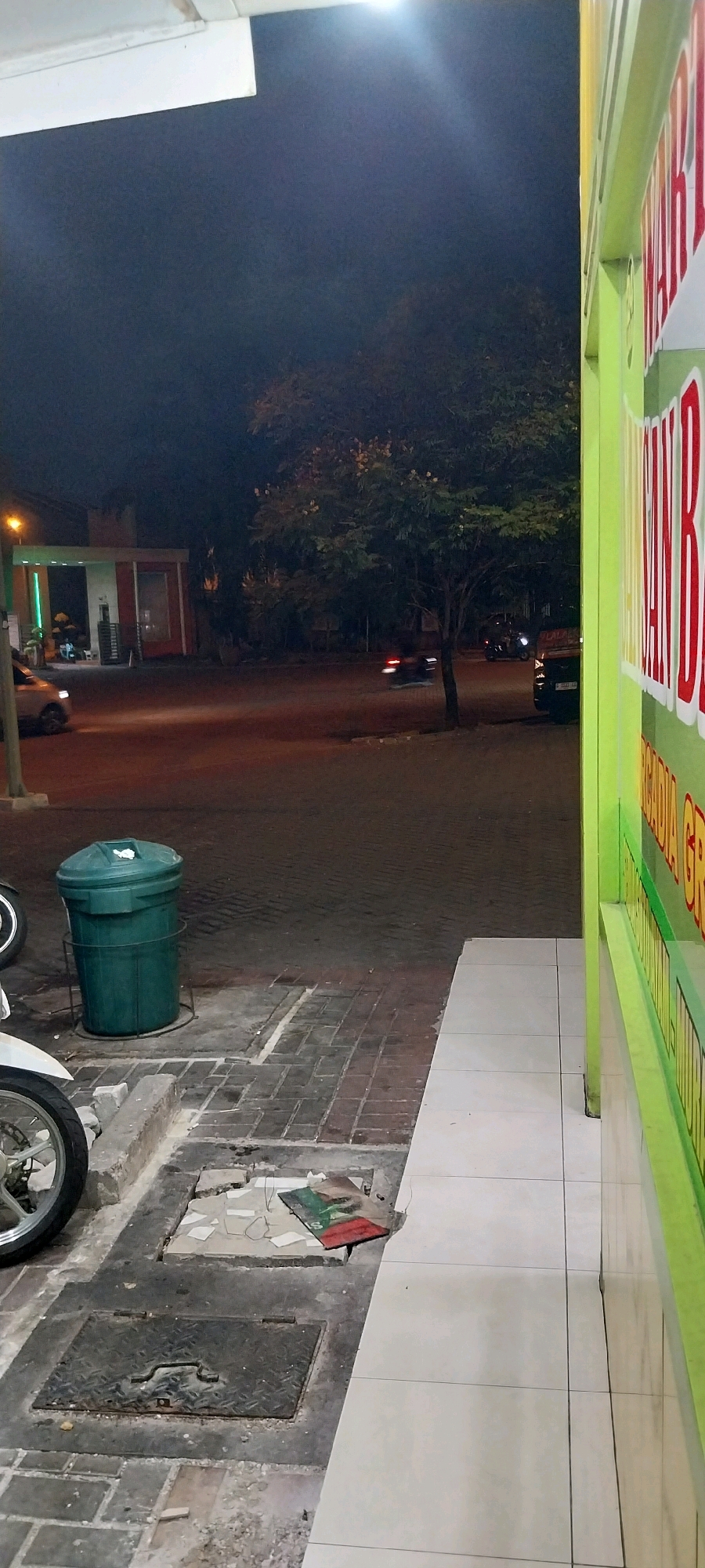 The image captures a street scene at night in Kelapa Dua, Indonesia. In the foreground, a white motorcycle is partially visible on the left, next to a large green trash can. The ground is paved with interlocking bricks and concrete. A metal manhole cover is present. To the right, the exterior of a building with a bright green facade and large, red lettering on it is visible, suggesting a commercial establishment. The background shows a street with a few cars and a motorcycle in motion, their headlights and taillights creating streaks of light, indicating movement. A building with an awning and some vegetation is also visible in the distance. The overall atmosphere is dimly lit, typical of a night-time urban environment.