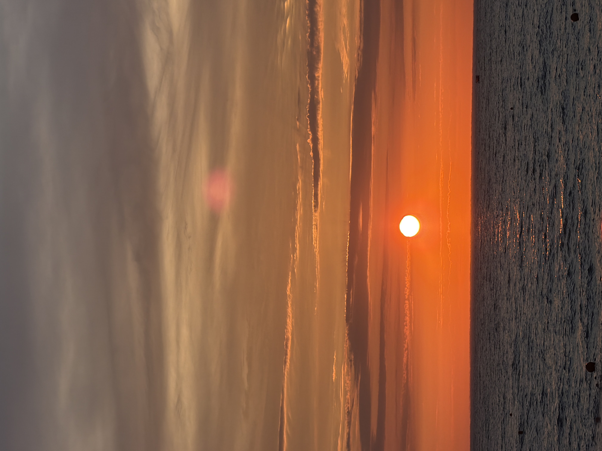 This is a vertical, full-frame photograph of a sunset over the sea. The sun, a bright orange orb, is low on the horizon, casting a warm glow across the sky and water. Wispy clouds streak across the sky, painted in shades of orange, pink, and gray. The sea is dark and choppy, reflecting the colors of the sunset. The overall mood is peaceful and serene, evoking the beauty of nature's spectacle. The image appears to have been taken from a beach or a coastal area. There are no people or man-made objects visible in the scene, except for a possible small, distant structure visible in the water on the right side of the frame. There is no visible text in the image. The time of day is sunset. The weather appears clear with some cloud cover. Given the location context, this scene is likely depicting a sunset over the Black Sea in Sochi, Russia.