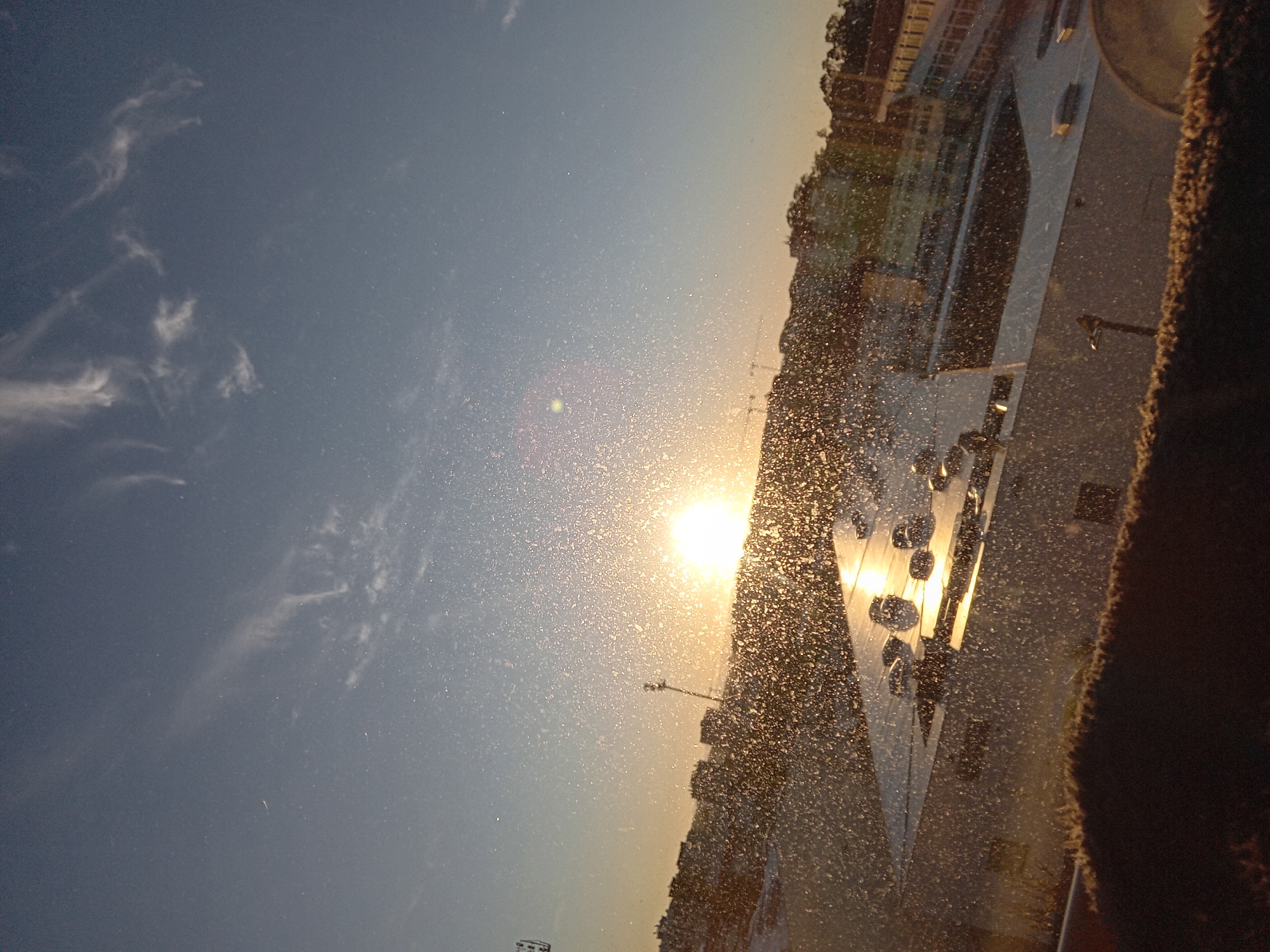 The image captures a scene at sunset with a water fountain as the central subject. The sun is positioned low on the horizon, casting a bright glare and causing the water droplets from the fountain to sparkle. In the background, buildings can be seen silhouetted against the evening sky. The camera appears to be positioned close to the fountain, as droplets are visible in the foreground, giving the impression of looking through a curtain of water. The overall scene conveys a sense of tranquility as the sun sets over the city.