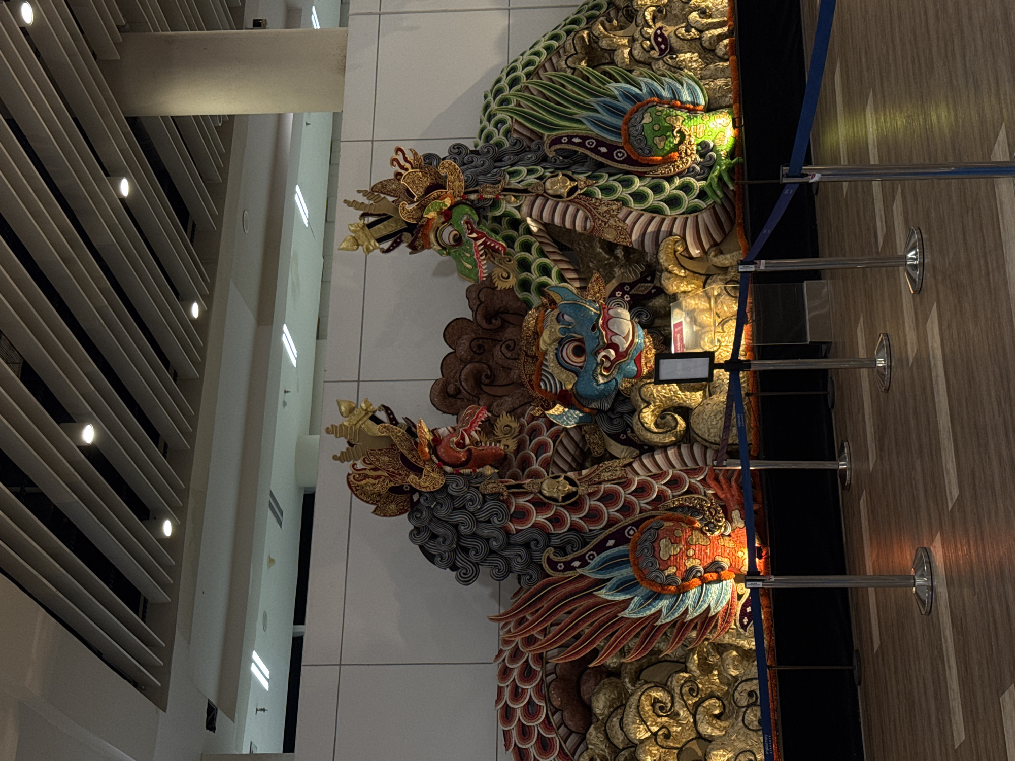A large, ornate, and colorful multi-headed dragon sculpture adorns a vertical wall in an indoor setting, likely a public space such as an airport, museum, or exhibition hall. The image is taken at an angle, capturing the dragon from its upper-right side.
The dragon sculpture itself is highly detailed, featuring vibrant hues of gold, green, blue, red, and brown, with intricate patterns forming scales, swirling clouds, and mythical embellishments. At least three distinct dragon heads are visible, each with prominent eyes, horns, and open mouths, facing towards the left side of the frame. One head, near the top, is primarily green and gold, while another below it features blue and reddish tones. A third head, further down, is dominated by red and gold. The craftsmanship suggests a rich cultural artistry, possibly East or Southeast Asian.
To the right of the dragon display, a light-colored wooden floor is visible, upon which several silver stanchions with blue retractable belts are set up, forming a barrier that suggests an area for viewing or crowd control. Artificial lighting illuminates parts of the dragon, highlighting its golden elements and vibrant colors.
On the left side of the image, architectural elements are visible, including a ceiling or upper wall structure composed of parallel white or light-colored slats or beams, interspersed with recessed circular lights that emit a soft glow. The overall lighting is artificial, indicative of an indoor environment. There are no people visible, and no specific activity is taking place other than the display of the artwork. A small white sign with black text is present on a black stand near the bottom right of the dragon, but the text is illegible.