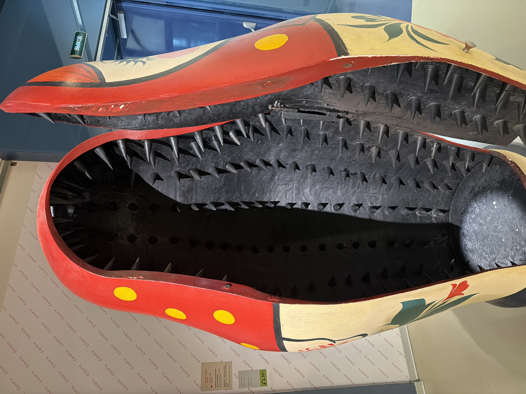 This is a photograph of a painted wooden trap with large teeth on display at the Erarta Museum of Contemporary Art in Saint Petersburg, Russia. The trap is painted in a style reminiscent of traditional Russian folk art, featuring red, yellow, and cream colors with floral designs. The interior of the trap is black, and the teeth are numerous and sharp. The trap is displayed on a white surface with repeated "ERARTA" lettering and is positioned near a blue-colored wall with a sign labeled "ВЫХОД". A small information card is visible near the trap.