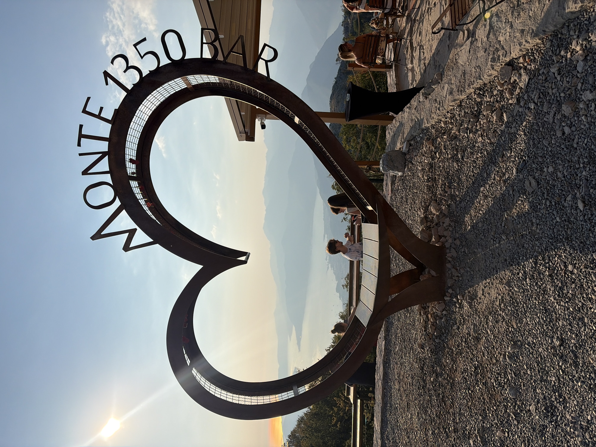 The image shows a heart-shaped sculpture with the text "MONTE 1350 B..." visible on its upper curve. Two people, a child and an adult, are seated at a table within the sculpture. In the background, a body of water is visible, surrounded by mountains. The scene appears to be a viewpoint or a restaurant, likely located at Monte 1350 in the vicinity of Kuk, Montenegro, given the text on the sculpture and the mountainous landscape. The sun is setting, casting a warm glow over the scene. Several other people are seen in the background, some seated at tables and chairs. The ground is covered in gravel.