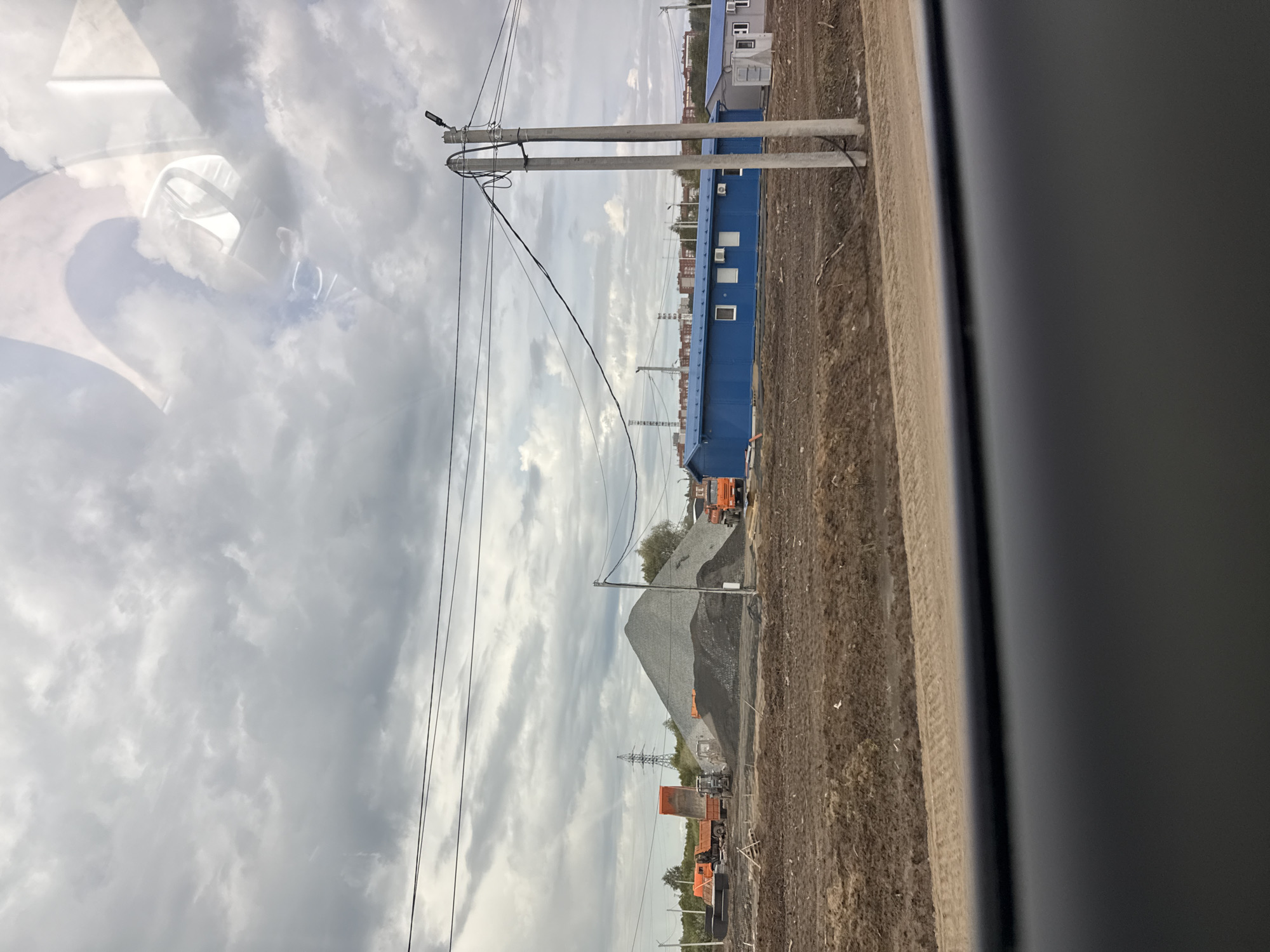 The image, taken from inside a vehicle and showing a reflection of its interior (dashboard, steering wheel, seats) on a window in the upper left, depicts an industrial or construction site under an overcast sky.
The scene is dominated by infrastructural elements and building materials. Several concrete utility poles with numerous power and communication lines stretch across the midground from left to right. To the right, a long, low blue building with multiple white windows and what appear to be air conditioning units is visible, possibly serving as an office or temporary structure.
Further back, large conical piles of grey and black crushed stone or gravel are prominent. Several orange dump trucks are positioned around these material piles, some appearing to be in use or waiting, with one seemingly loaded. In the distance, an electrical transmission tower stands against the cloudy horizon, and other smaller buildings or structures are faintly visible. The ground throughout the visible area is largely undeveloped, consisting of disturbed earth and dirt.
The sky is heavily clouded, suggesting an overcast day with no direct sunlight. No people are clearly visible, but the presence of machinery and prepared materials indicates ongoing or recent industrial activity. There is no discernible text in the image. The setting appears to be an undeveloped area undergoing construction or serving as a material depot, consistent with an industrial or infrastructure project in Kabanikha, Russia.