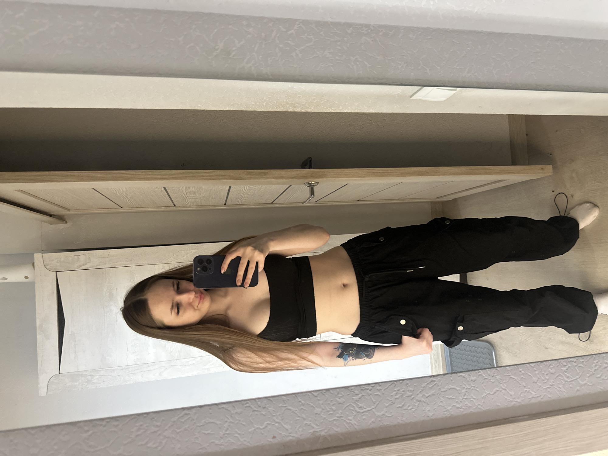 A young woman is visible in a reflected view, seemingly taking a self-portrait in what appears to be an indoor setting. She is wearing a black crop top and baggy black pants. Her long brown hair is visible. She is holding a smartphone in her hand. The reflection shows a wall and part of a cabinet or wardrobe. The location is unknown.