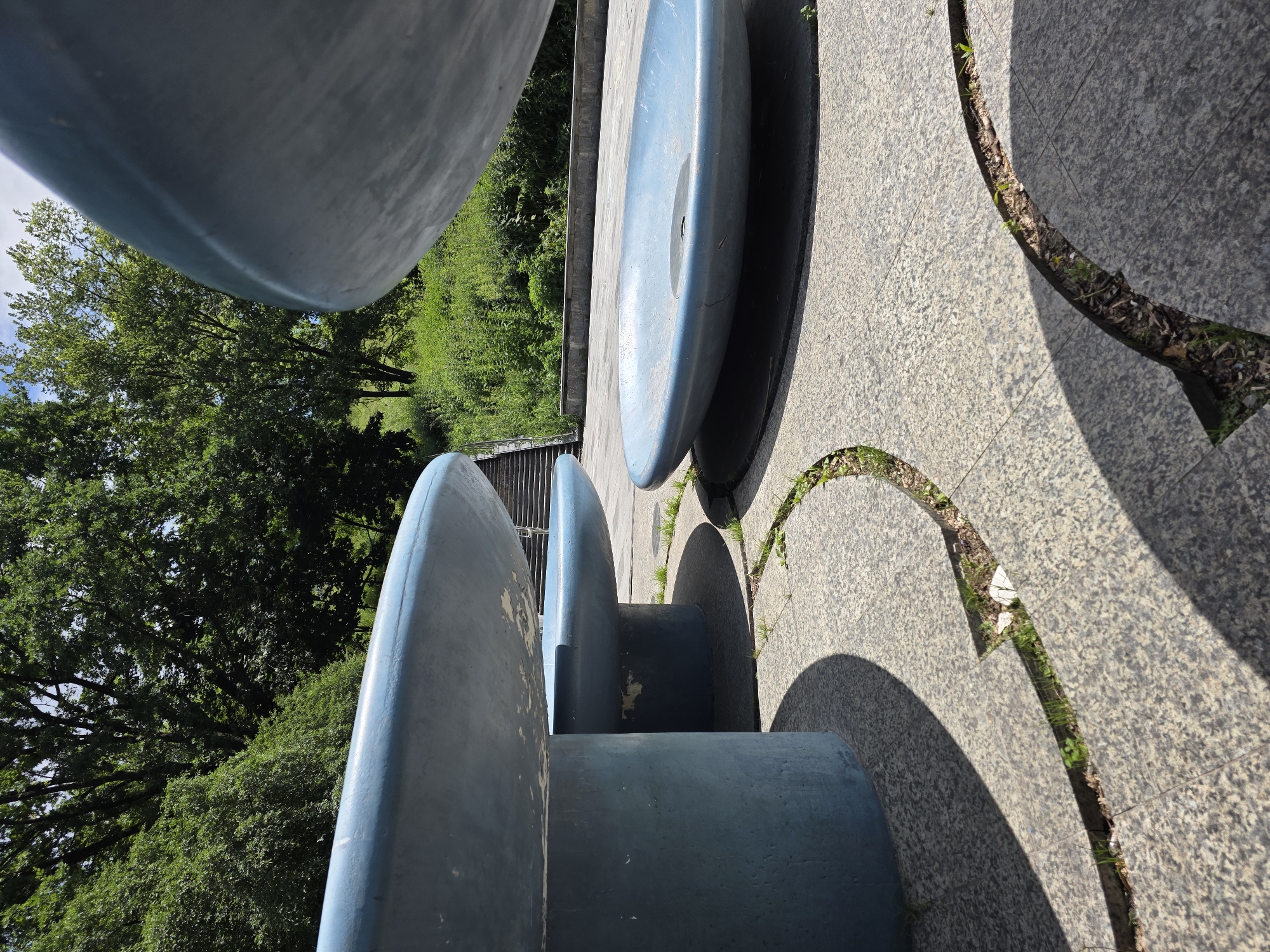 The image captures a section of the Memorial to the Victims of the Holodomor in Kyiv, Ukraine. In the foreground, there are several large, blue, bowl-shaped concrete structures, likely part of a water feature or sculptural element. These structures are positioned on a grey, speckled paved surface with curved channels filled with dirt and sparse vegetation. The lighting suggests it is a sunny day, casting strong shadows from the concrete forms. In the background, a lush green hillside is visible, topped with mature trees. To the left, a set of concrete stairs ascends the slope. The overall scene is serene and monumental, evoking a sense of historical reflection.