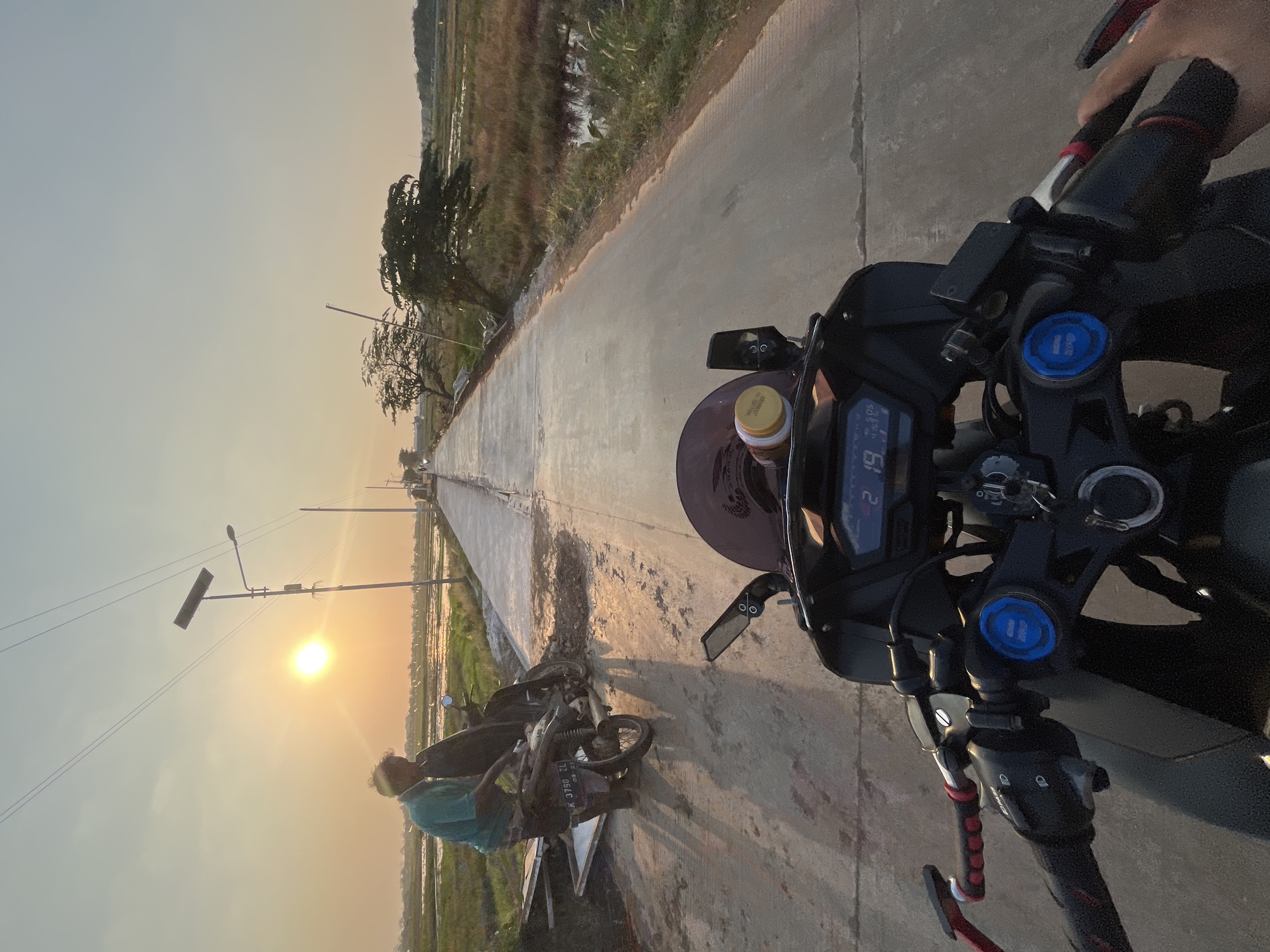 The image depicts a first-person perspective from a motorcycle on a paved road in Pecangaan, Indonesia, at sunset. The road stretches into the distance, flanked by rice paddies on either side. In the mid-ground, a man in a teal shirt is standing next to a parked motorcycle, appearing to interact with it. Solar-powered streetlights line the road. The sky is a soft orange and yellow due to the setting sun, casting long shadows. The motorcycle in the foreground shows its handlebars, digital display, and a jar secured to the windshield. The environment suggests a rural or semi-rural setting within Pecangaan.