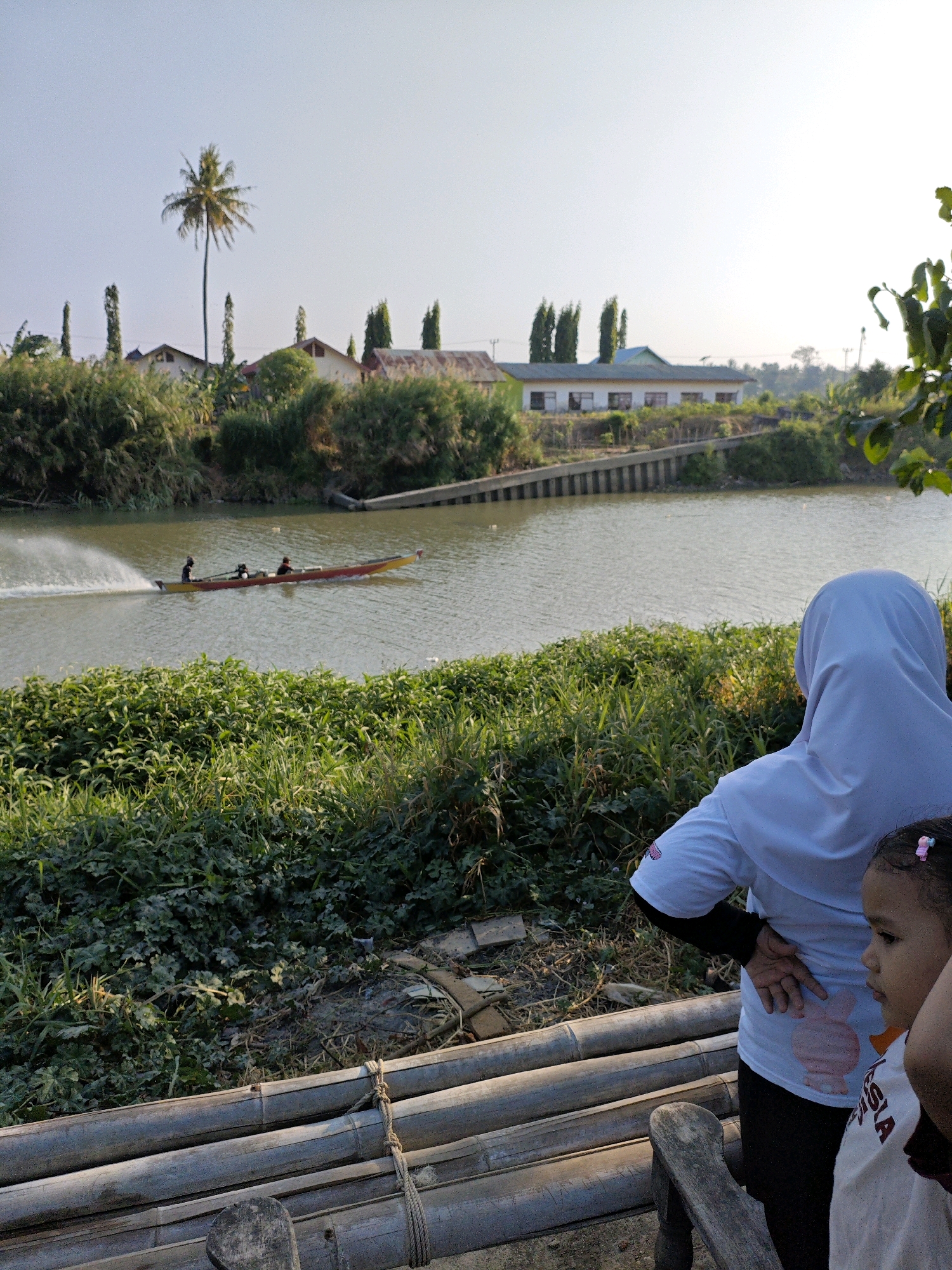 In the city of Padduppa, Indonesia, an outdoor scene unfolds by a wide river. A long, narrow boat, colored red and yellow, is speeding down the water, leaving a white wake. Three people are visible in the boat. On the right bank of the river, a woman wearing a white hijab and a white shirt is looking towards the water. A young child is standing next to her, also looking in the same direction. In the foreground, stacked bamboo poles, tied with rope, form a rustic barrier. Behind the river, lush green vegetation lines the banks, with trees and some buildings visible in the distance, suggesting a rural or semi-urban setting. The sky is clear and bright, indicating a sunny day.