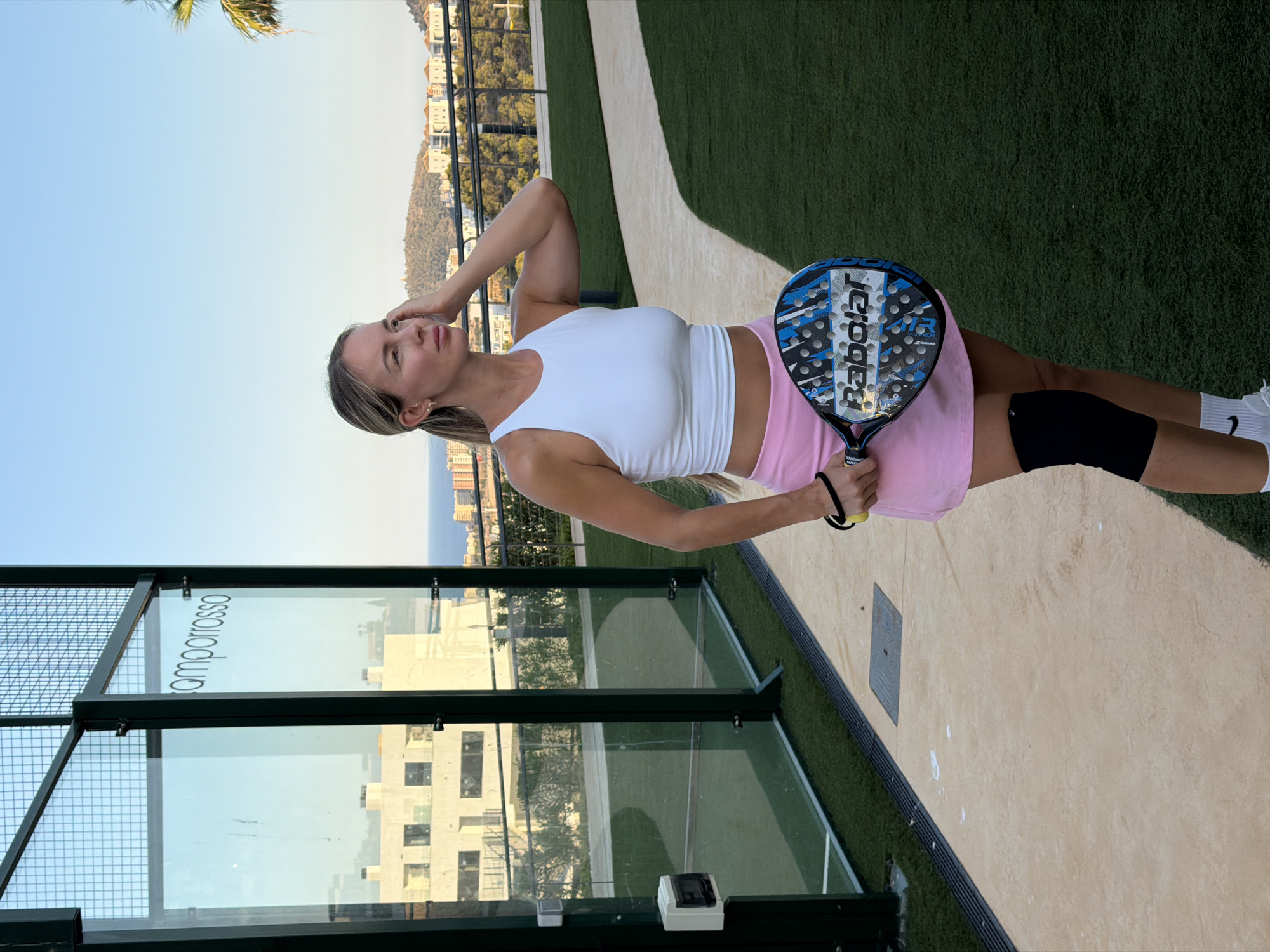 A woman with blonde hair pulled into a ponytail stands on a paved walkway adjacent to a green astroturf court, holding a blue and black paddle tennis racket. She is wearing a white cropped tank top, a pink skirt, a black knee brace, and white athletic socks with sneakers. The court has glass walls with a metal frame. In the background, buildings and hills are visible under a clear blue sky. The image appears to have been taken in Finestrat, Spain, given the architectural style and landscape.