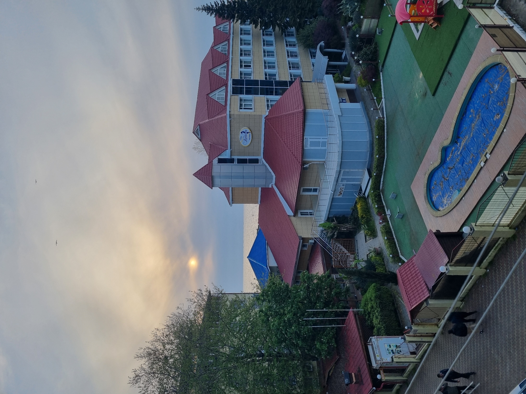 The image, taken in Sochi, Russia, captures an outdoor scene at a hotel or resort complex, likely during either sunrise or sunset, given the soft, diffused golden light. The view looks out towards the sea, visible in the background under a cloudy sky where the sun's glow creates a distinct halo effect. A bird is seen flying in the sky.
The prominent subject is a multi-story hotel building with yellow facades and distinctive red tiled roofs, featuring dormer windows. A large circular sign on the hotel reads "HOTEL" and "ЮЛИЯ" (Yuliya/Julia), with "РЕСТОРАН" (Restaurant) underneath. Adjacent to the main building is an outdoor recreational area. This area includes expansive green lawns, a kidney-shaped swimming pool covered with a blue protective tarp, and a children's playground equipped with a red and orange slide and climbing structure. Various trees and shrubs provide greenery around the property, and outdoor lighting fixtures are visible along pathways and around the pool area.
Closer to the foreground, a paved area is visible where a few individuals are walking, appearing as dark silhouettes. The sea is situated beyond the hotel buildings, with another building featuring a blue roof visible closer to the coastline. Metal railings and other architectural elements are present throughout the complex. A smaller white sign near the paved area is partially visible, showing some text, including what appears to be "HOTEL." The overall impression is that of a tranquil resort setting.