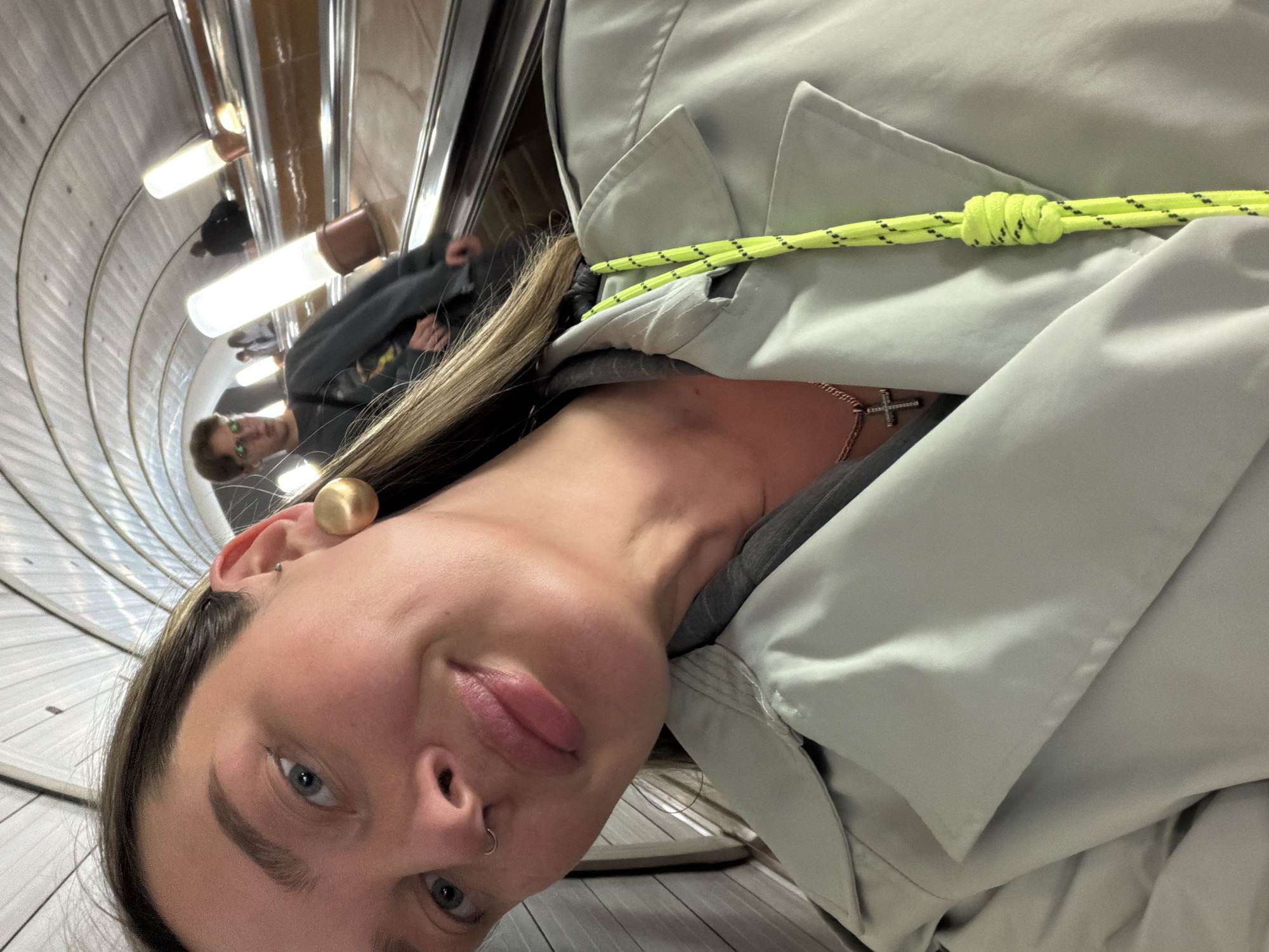 A selfie of a woman with blonde hair and blue eyes is taken on a descending escalator. She is wearing a gold sphere earring, a nose ring, and a silver chain with a cross. A light green jacket with a neon green and black rope detail is also visible. Behind her on the escalator is a man in a grey sweater and another figure further back. The escalator's structure is a tunnel shape with repeating metal lines and illuminated lights along the walls, indicating an underground location. This location is likely the Moscow Metro due to its unique architectural style.