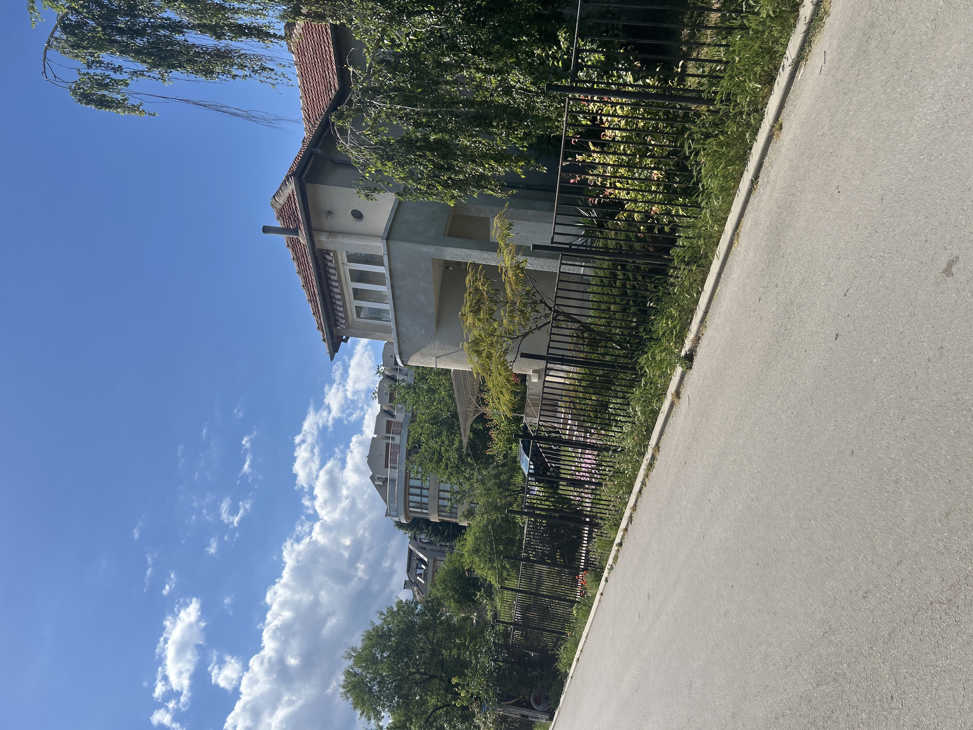 The image displays a street scene in Varna, Bulgaria, on a clear, sunny day with scattered clouds. The perspective is from a sidewalk looking towards a row of houses and lush green trees. The houses, with their tiled roofs and varied architectural styles, are partially obscured by the foliage. A black metal fence runs along the property line, separating the houses from the sidewalk. The sidewalk itself is paved with asphalt. The sunlight creates strong shadows, suggesting it is mid-morning or afternoon. The overall impression is one of a peaceful, residential neighborhood.
