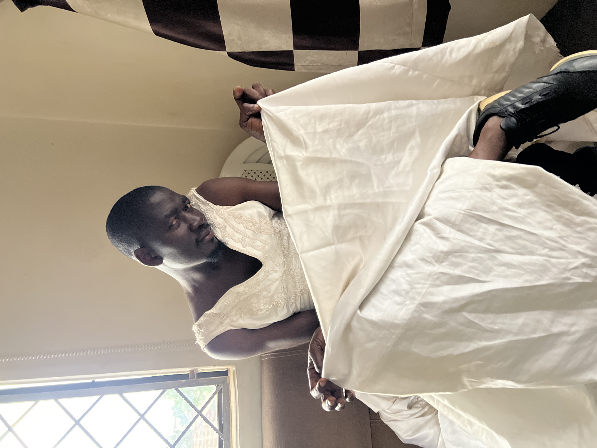 In Jos, Nigeria, a Black man is seated indoors, wearing a cream-colored sleeveless dress with intricate beading on the bodice. He is holding the fabric of the dress, which is a lighter cream color and appears to be satin, in his hands and looking down at it. He has short dark hair and is wearing black athletic shoes with yellow accents. Behind him, a window with a diamond pattern pane is visible on the left, allowing natural light to enter the room. To the right, a curtain with a checkerboard pattern of dark brown and white hangs. The man is seated on a brown couch.