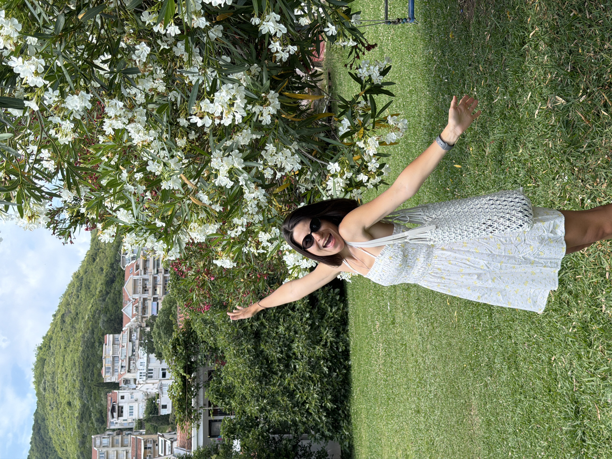 A young woman with short dark hair and sunglasses stands in a grassy yard with her right arm raised and her left arm extended, her mouth open in a laugh. She is wearing a white floral sundress and a white crocheted bag hangs from her left shoulder. Behind her, a lush green hillside is covered with apartment buildings and trees. A large white flowering bush is to her right. The location appears to be a residential area in Budva, Montenegro, likely on a warm, sunny day.