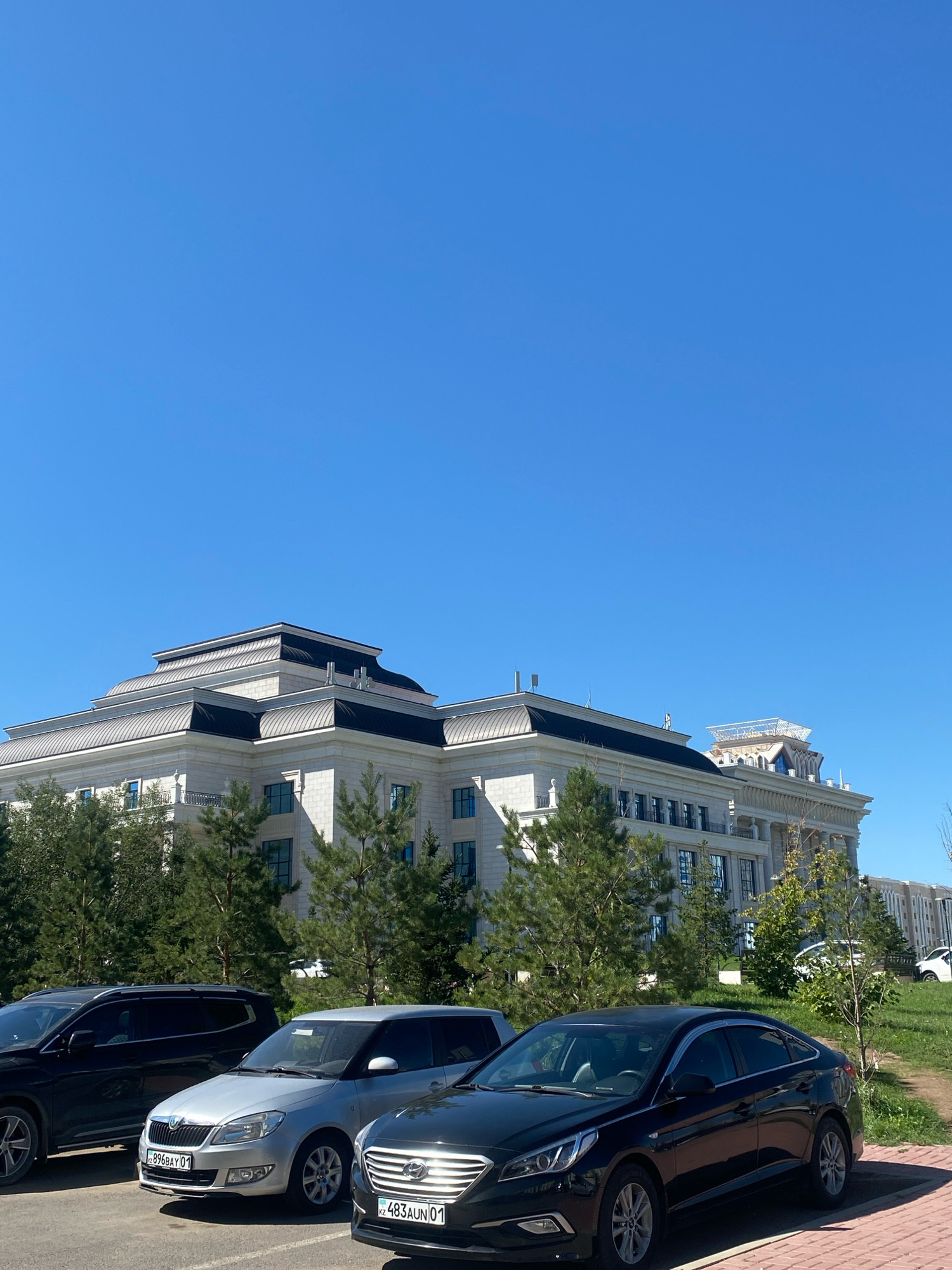 In Astana, Kazakhstan, a large, modern building with a multi-tiered, dark metal roof and light-colored stone facade stands under a clear blue sky. Evergreen trees line the front of the building, and a parking lot in front of it contains several vehicles. Three cars are visible in the foreground: a black SUV, a silver hatchback, and a black sedan. The silver hatchback has a license plate that reads "KZ 896BAY 01". The black sedan has a license plate that reads "KZ 483AUN 01". The environment is clean and orderly, suggesting a well-maintained public or corporate space.