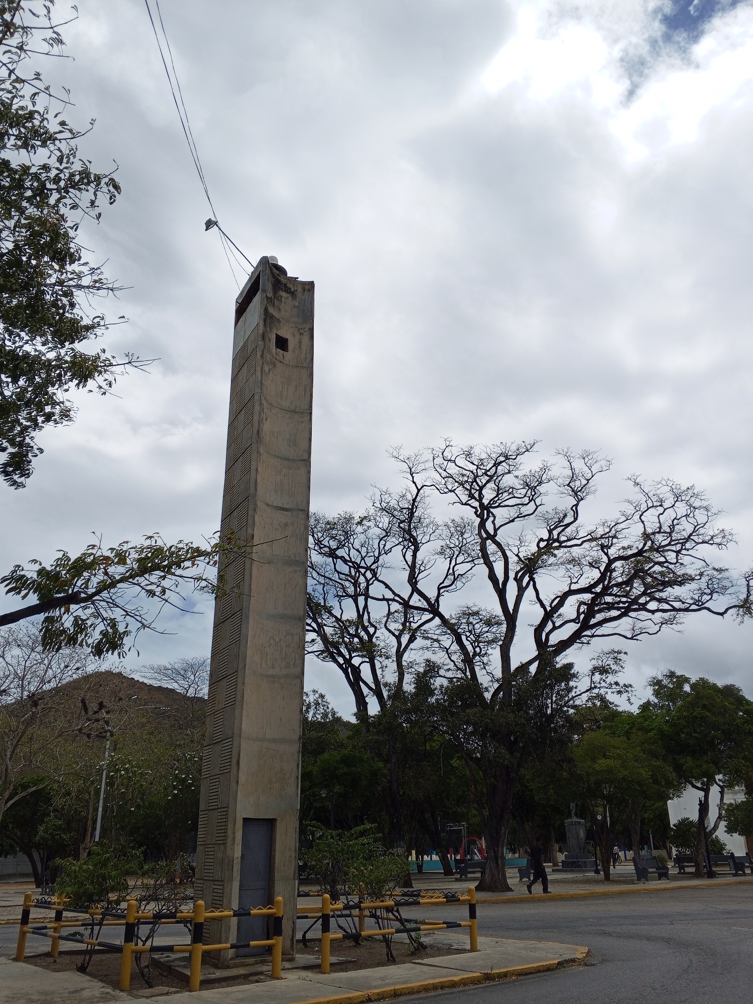 A tall, concrete structure stands prominently in a park setting. The structure features a small window near the top and a door at the base, enclosed by a low yellow and black barrier. Bare-branched trees stand behind it, contrasting with the overcast sky. In the background, other park elements like benches, trees, a statue, and indistinct figures can be seen. Power lines extend into the sky from the upper left of the structure. The image is taken in a city of Unknown, Unknown.