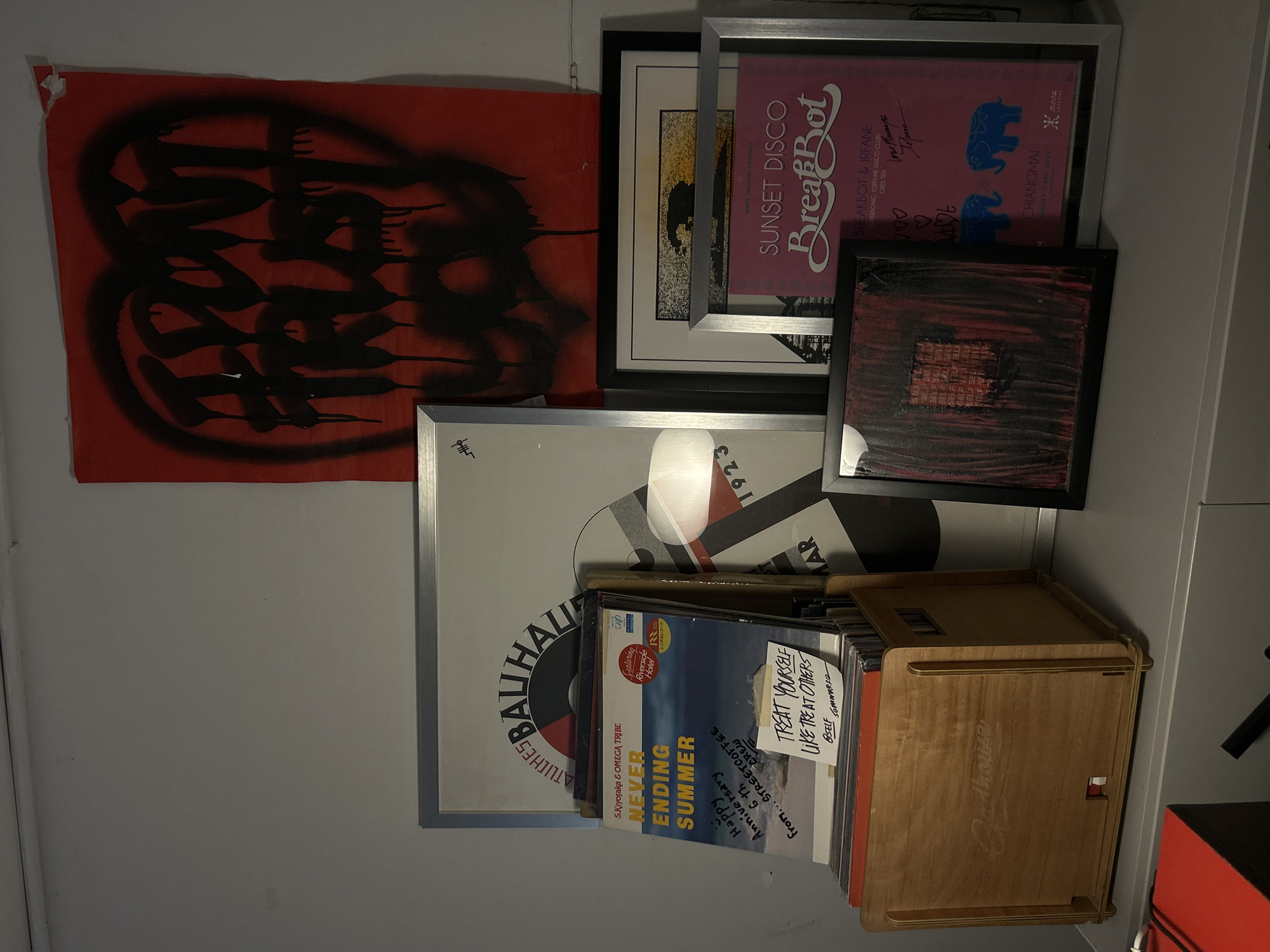 The image shows a shelf with several vinyl records and framed art. On the right side of the image, there is a red poster with black spray paint that says "I DON'T TRUST". Below it, there are two framed pieces of art. The top one is a black and white image with some yellow. The bottom one is a pink poster with the text "SUNSET DISCO BreakBot" in stylized font, and two blue elephants. There are also handwritten messages and drawings on this poster, including "love always".
To the left of the framed art, there is a stack of vinyl records. The top record cover has the title "NEVER ENDING SUMMER" and a handwritten message that reads "Happy Anniversary 6th From... STREETCOFFEE". Another note on this record says "TREAT YOURSELF LIKE TREAT OTHERS". These records are in a wooden crate with the embossed text "arthouse".
Behind the vinyl records, there is a large framed poster with text that appears to be "BAUHAUS".
The lighting suggests an indoor environment, possibly a room or a display area. The overall aesthetic is somewhat eclectic, with a mix of graphic design, art, and personal touches.
There is no information in the image that indicates the location is in the city of Chiang Mai City Municipality, Thailand.