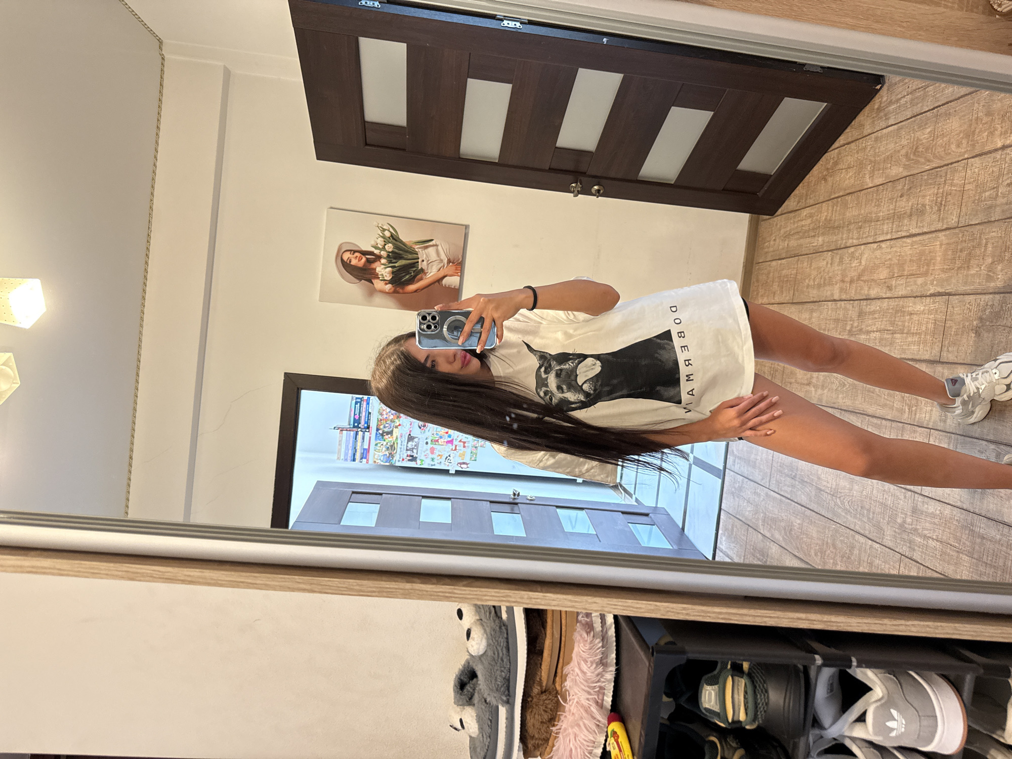 A young woman with long, dark hair stands in front of a full-length mirror, taking a selfie with her phone. She is wearing a white t-shirt with a black graphic of a Doberman and text, and black shorts. Her legs are bare and she is wearing white athletic shoes. In the mirror's reflection, a shoe rack with various shoes and slippers is visible to the left. A painting of a woman holding flowers hangs on the wall behind her. Two dark wooden doors are also visible in the reflection. The floor is made of light brown wooden planks. The environment appears to be an indoor residential setting, likely an entryway or hallway, within a building in Chișinău, Moldova.