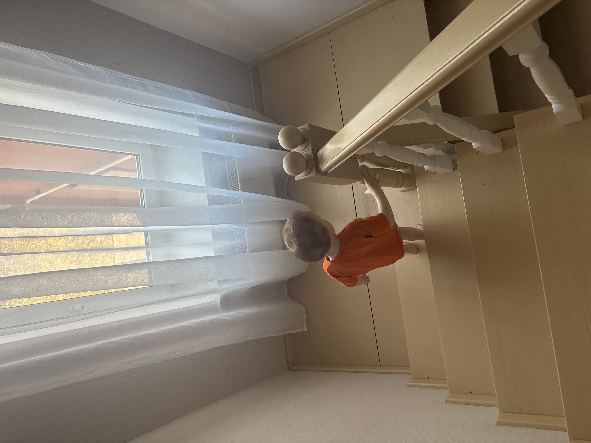 A high-angle shot depicts a young child wearing an orange shirt standing on a beige carpeted floor near the bottom of a wooden staircase. The child is reaching for the white balustrade of the staircase. A window with white blinds is situated nearby, covered with sheer white curtains. The photo was taken indoors. The location of the image is in Ферма, Russia.