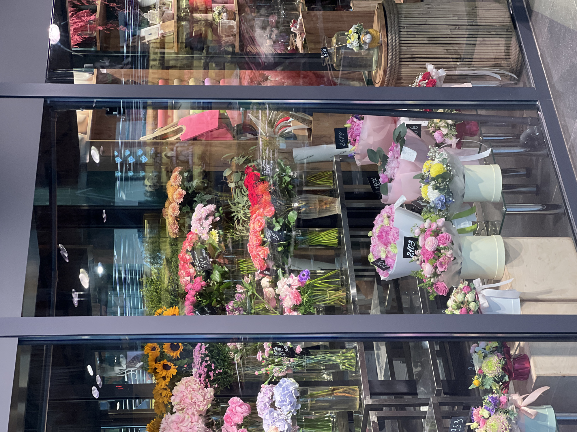 The image shows a storefront display of a flower shop. The display features a variety of bouquets and arrangements of fresh flowers in vases and boxes. The flowers are in various colors, including pink, yellow, red, and purple. There are also some decorative items visible in the background, such as rolls of wrapping paper and shelves stocked with vases. The storefront appears to be a large glass window, allowing a clear view of the merchandise inside. The lighting suggests it is daytime, and the reflection on the glass indicates an indoor setting. The overall impression is of a well-organized and attractive display of flowers. The text on the price tags suggests the location is in Kyiv, Ukraine, with prices indicated in both local currency and potentially euros.
