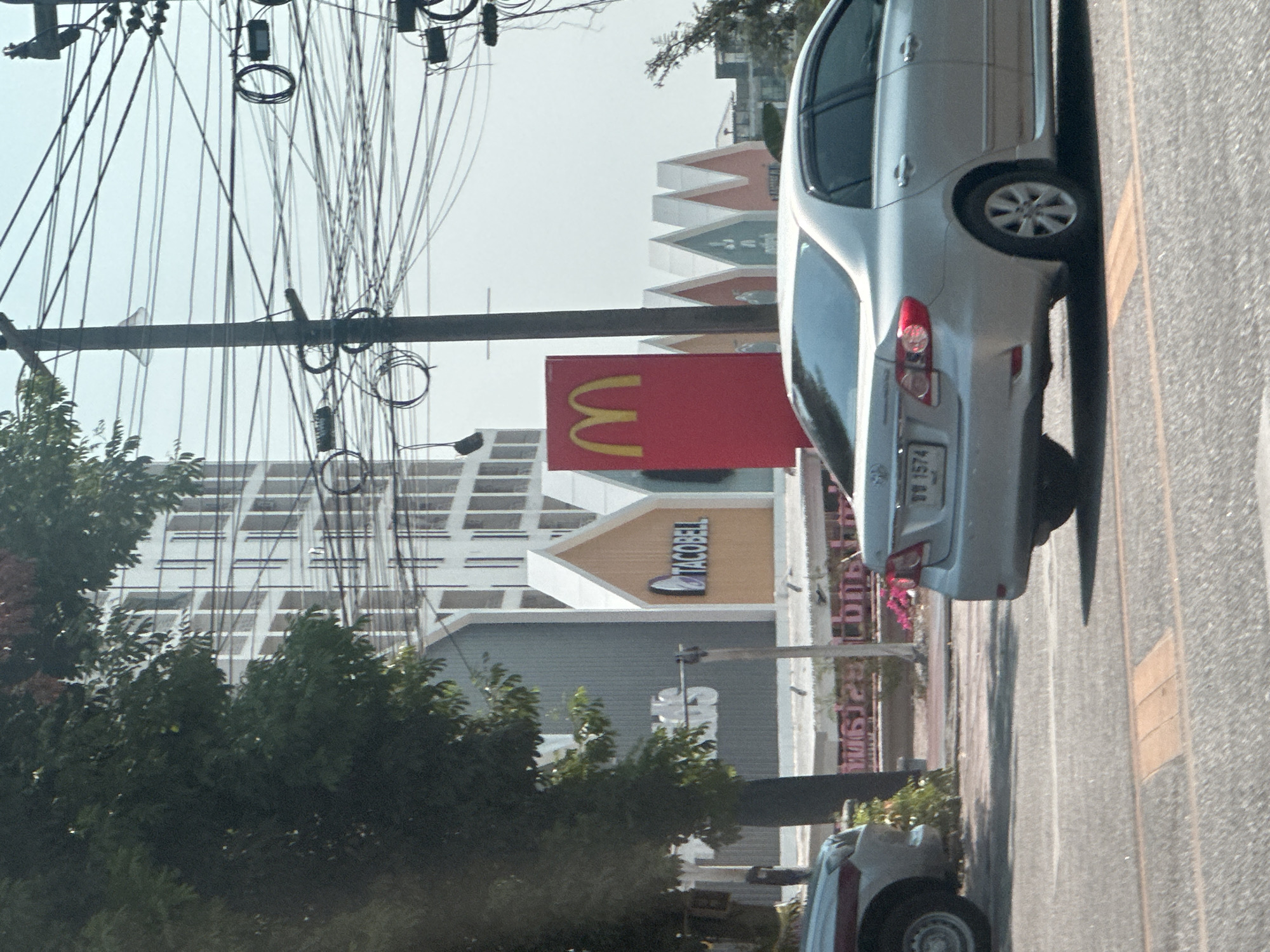 The image captures a street scene in Pattaya City, Thailand. A grey sedan occupies the right side of the frame, partially obscuring the view of buildings and commercial signage. A McDonald's sign with the iconic golden arches is visible, along with signage for Taco Bell and another establishment. Overhead power lines are densely strung across the sky. Trees and shrubs are visible, and another vehicle can be seen in the lower part of the frame.