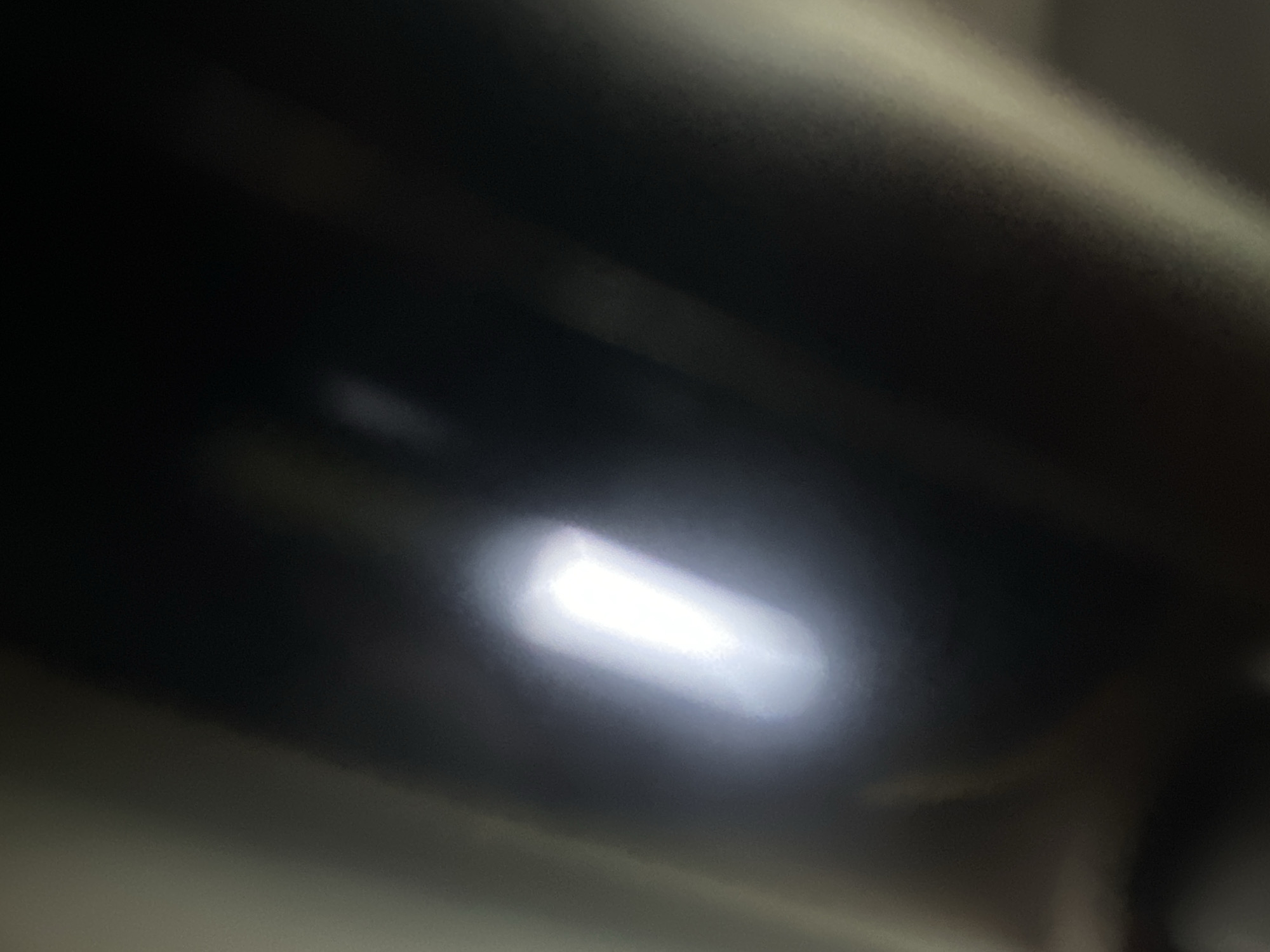 This is a close-up, abstract image dominated by dark, blurry shapes and a bright, elongated rectangular light source. The overall impression is of a dark, possibly metallic or highly polished surface reflecting light. The light source is intensely bright and appears to be a LED strip or a similar linear light, casting a strong glow that creates a halo effect around its edges. The surrounding darkness suggests an interior setting or a dimly lit environment where this light is the primary focal point. There are no discernible people, specific objects, or clear scene elements, making it difficult to ascertain the exact setting or activity. The focus is entirely on the interplay of light and shadow on an abstract surface. There is no visible text in the image. The location context of Belgrade, Serbia, does not offer any specific visual cues within this abstract representation.