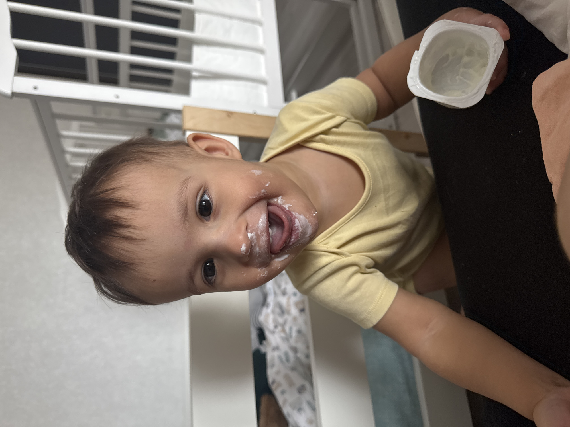 A baby in a yellow shirt sits near a white crib in what appears to be an indoor environment. The baby's face and tongue are smeared with a white substance, possibly yogurt. The baby is holding a small, empty, white container. Part of a person's arm is visible, holding the container, likely an adult caregiver. The image suggests the baby has been eating, and the environment seems to be a home, potentially in the city of Chaikovsky, Russia, as requested, although no specific visual markers confirm the location.
