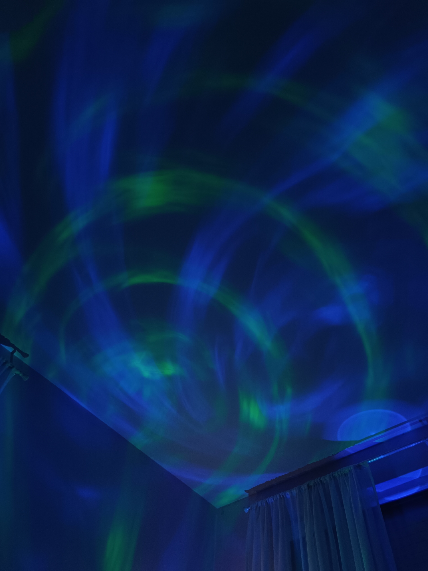 The image depicts an indoor scene with a corner of a room illuminated by swirling green and blue lights, likely from a projector. The lights are projected onto the ceiling and upper walls. The lower part of the image shows a white curtain hanging from a rod. The location in the city of Cherkovitsa, Bulgaria is not discernible from the image. There are no people, subjects, objects, actions, or interactions in the image. The environment is an indoor room.