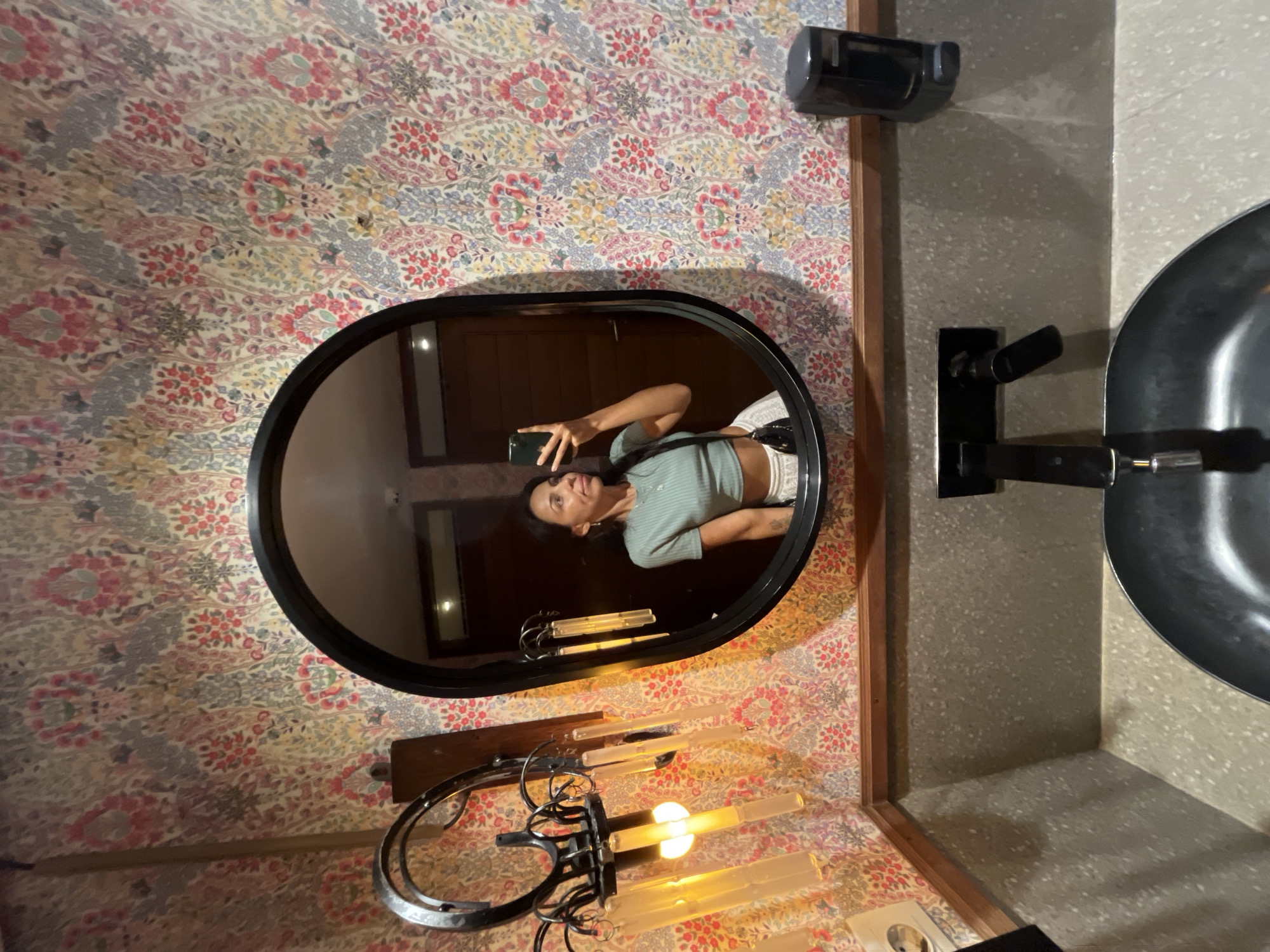 A selfie reflected in an oval mirror shows a woman holding a phone, wearing a light green top and white shorts, with tattoos visible on her abdomen. The mirror is hung on a wall covered with floral patterned wallpaper. Below the mirror is a sconce with multiple light tubes, some illuminated. To the right of the mirror, a black metal faucet is attached to a grey speckled wall near a dark metal washbasin. A black object, possibly a soap dispenser, is mounted to the wall above the mirror. The location appears to be a bathroom in Denpasar, Indonesia, suggested by the style of wallpaper and fixture.
