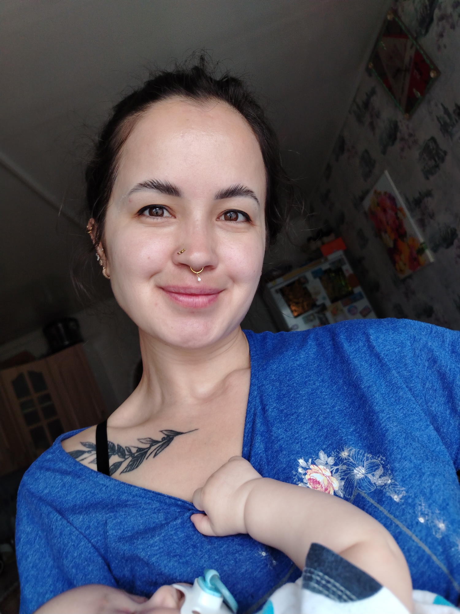 A close-up, slightly high-angle shot shows a woman holding a baby. The woman is fair-skinned with brown eyes and dark hair pulled back. She has multiple piercings in her ear and nose. She is wearing a blue t-shirt, revealing a black bra strap and a tattoo of a leafy branch on her upper chest. The baby, partially visible, is wrapped in a blanket with white, blue, and gray patterns. The woman is smiling slightly. The background includes a kitchen cabinet and a wall with floral-patterned wallpaper and framed artwork. The location is Unknown, Unknown.