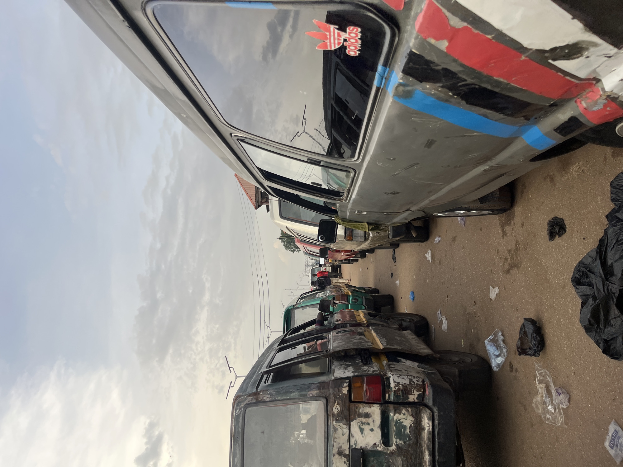 This media file captures a street scene in Bukuru, Nigeria, likely during the daytime with overcast skies. The primary subjects are a line of worn and weathered minibusses, commonly known as danfo buses, parked on a paved road. The danfos are in various states of disrepair, with faded paint, rust, and visible damage. Some have makeshift repairs and decorations, such as the red, black, and blue striped tape on the side of the nearest bus, and an Adidas logo sticker on its window.
The setting appears to be a busy area, possibly a bus park or a marketplace, given the presence of multiple vehicles lined up. The ground is littered with plastic bags and other debris, suggesting a less than pristine environment. The sky is cloudy, indicating either early morning or late afternoon, or simply a generally overcast day. There are no visible people in the foreground, but the interiors of some buses suggest they may be occupied or recently used. The overall impression is one of a functional but somewhat neglected urban transportation hub, characteristic of many developing cities. The scene conveys a sense of daily life and the challenges of maintaining public transportation infrastructure.