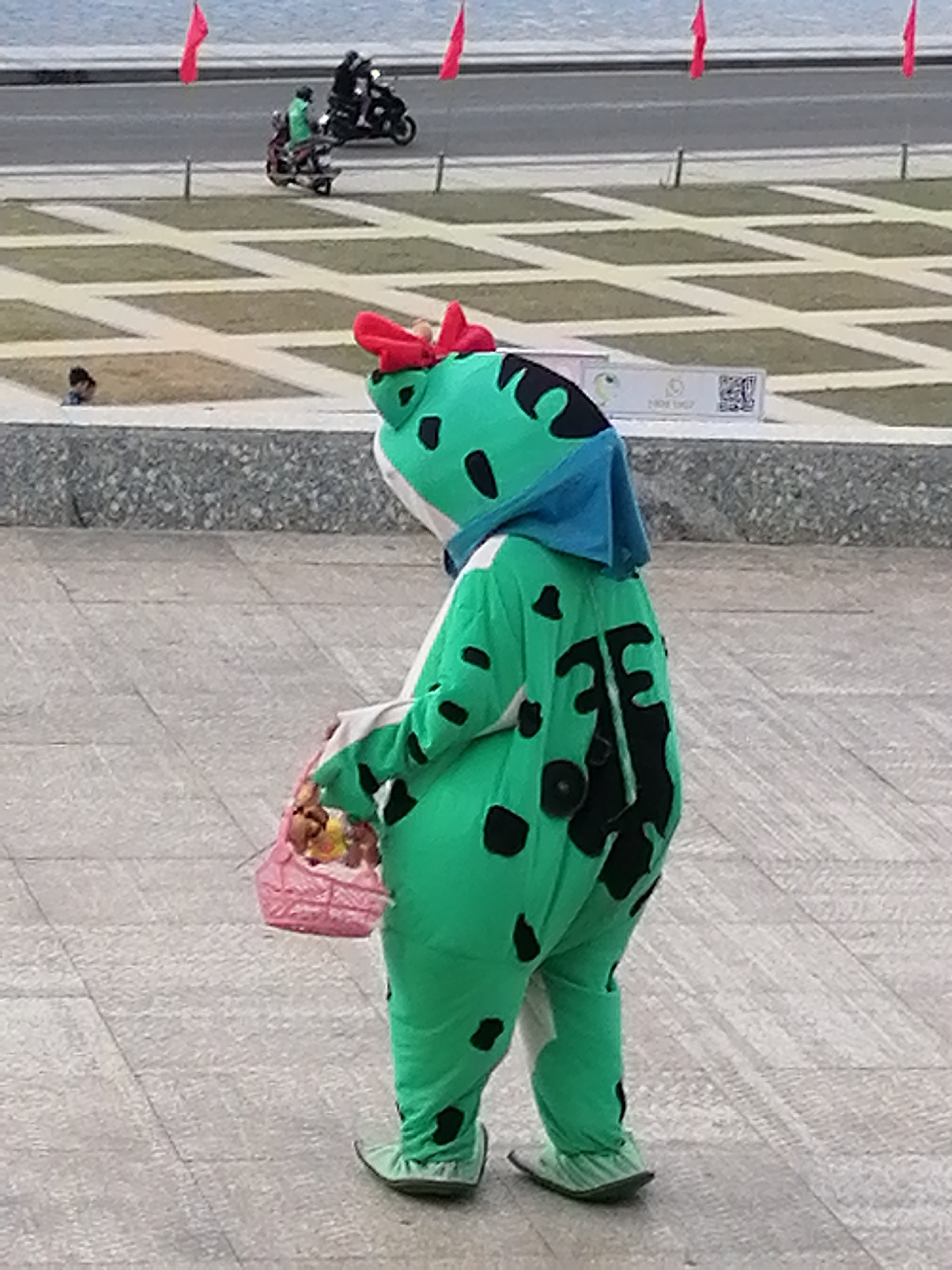 A person in a green cartoon character costume, with red accents on the head and black spots on the body, is holding a pink basket containing what appears to be food items. The costume has a blue hood. The person is standing on a paved walkway with a patterned background of light-colored squares. In the background, there is a road with a body of water beyond it. Several red flags are visible on poles along the roadside. Two people are riding on motorcycles on the road, and there is a grassy area with a geometric pattern between the walkway and the road. A small child is visible sitting on the grass in the distance. The scene appears to be outdoors, possibly in a public area such as a park or promenade in Da Lat, Vietnam, indicated by the presence of red flags which are a common feature in Vietnamese public spaces and the architecture of the paving.